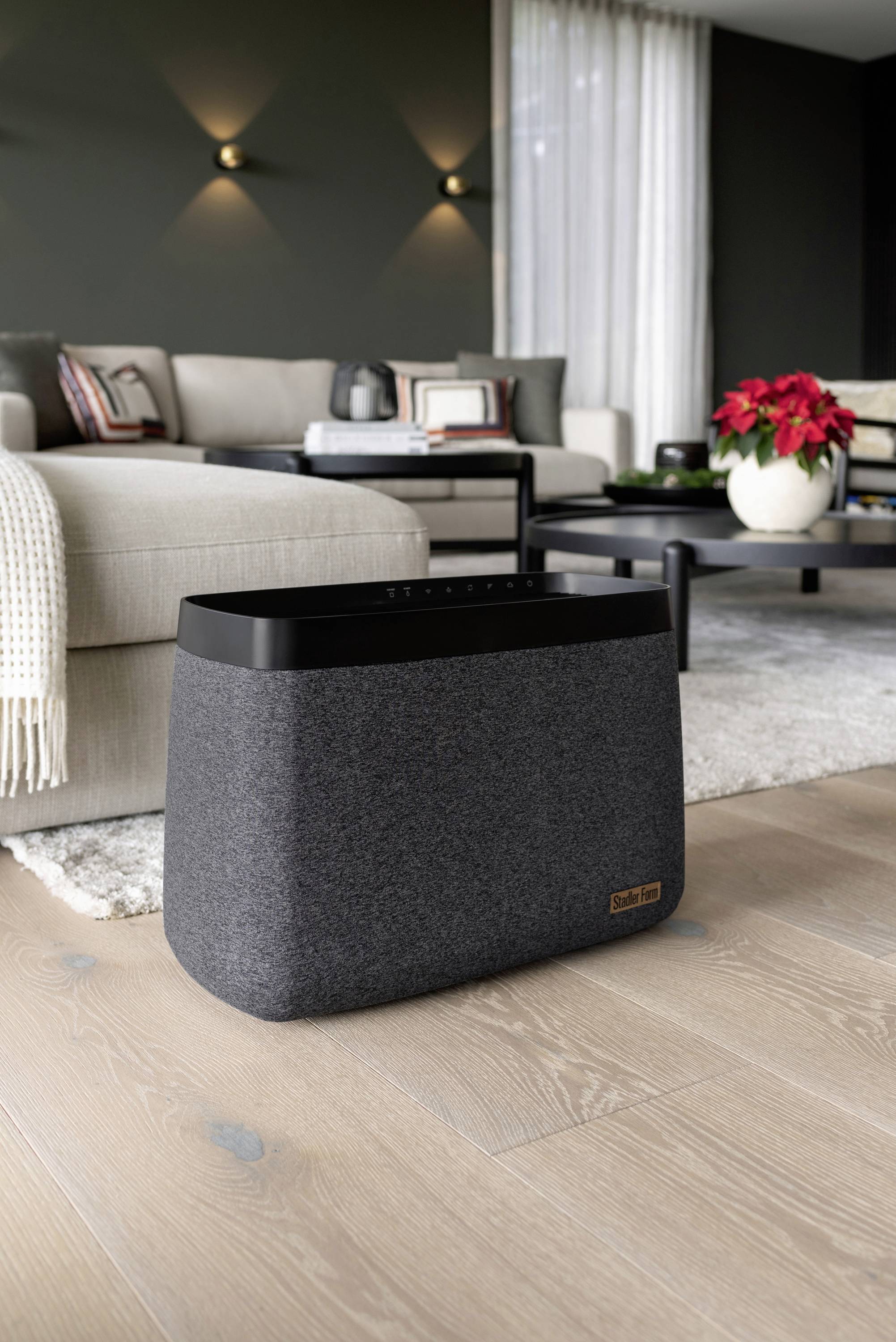 A grey speaker sits on a wooden floor in a modern living room. In the background, a beige sofa and a table with flowers are visible.