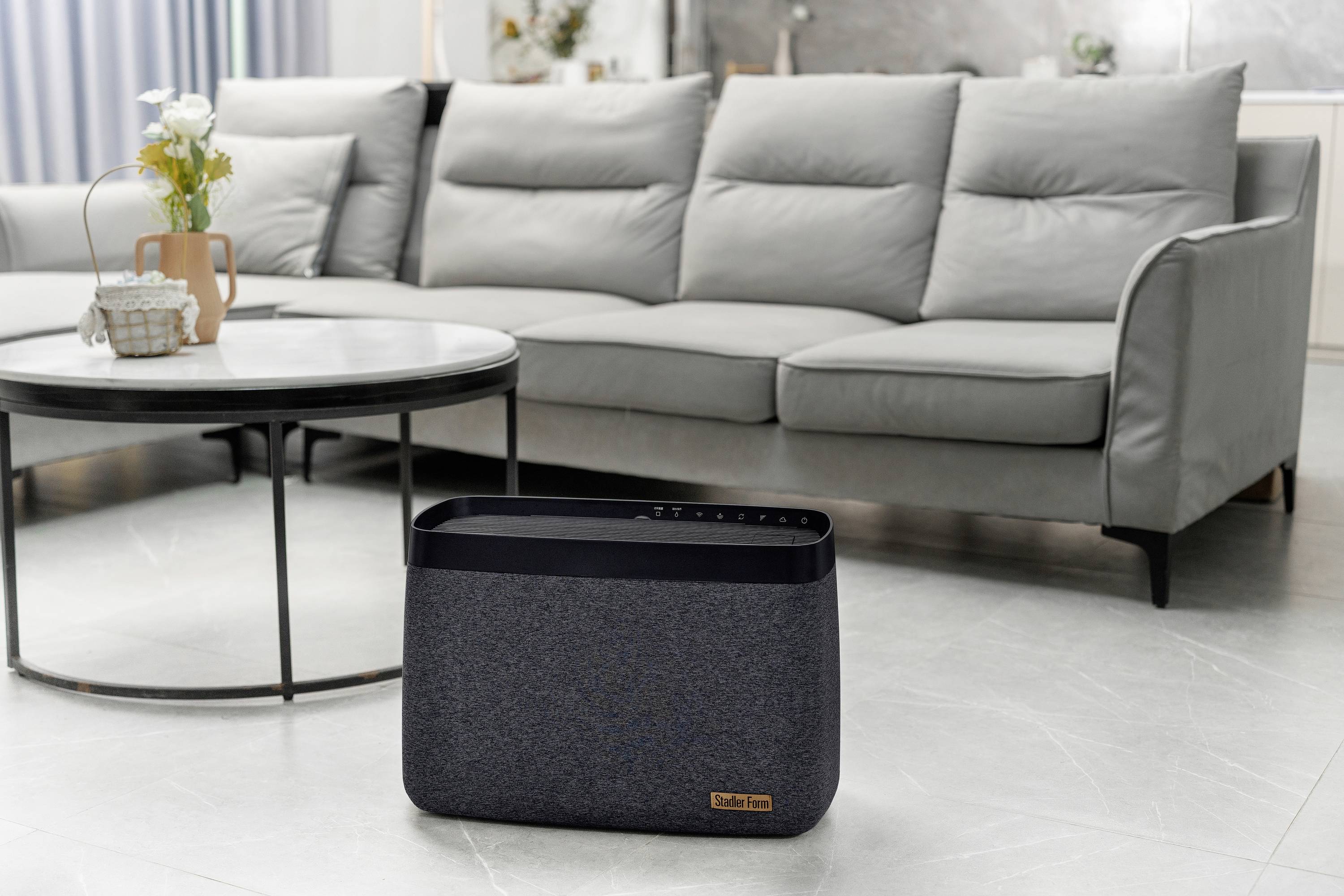 A grey air purifier stands in a modern living room with a grey sofa and a round coffee table in the background.