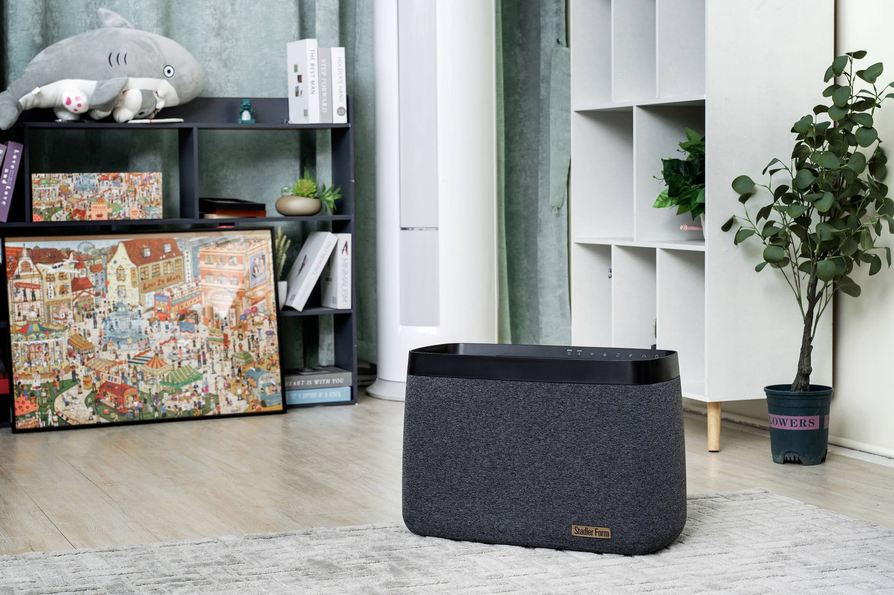 A living room with a grey air purifier on the floor, a shelf with plants, and a colourful puzzle canvas painting on the wall.
