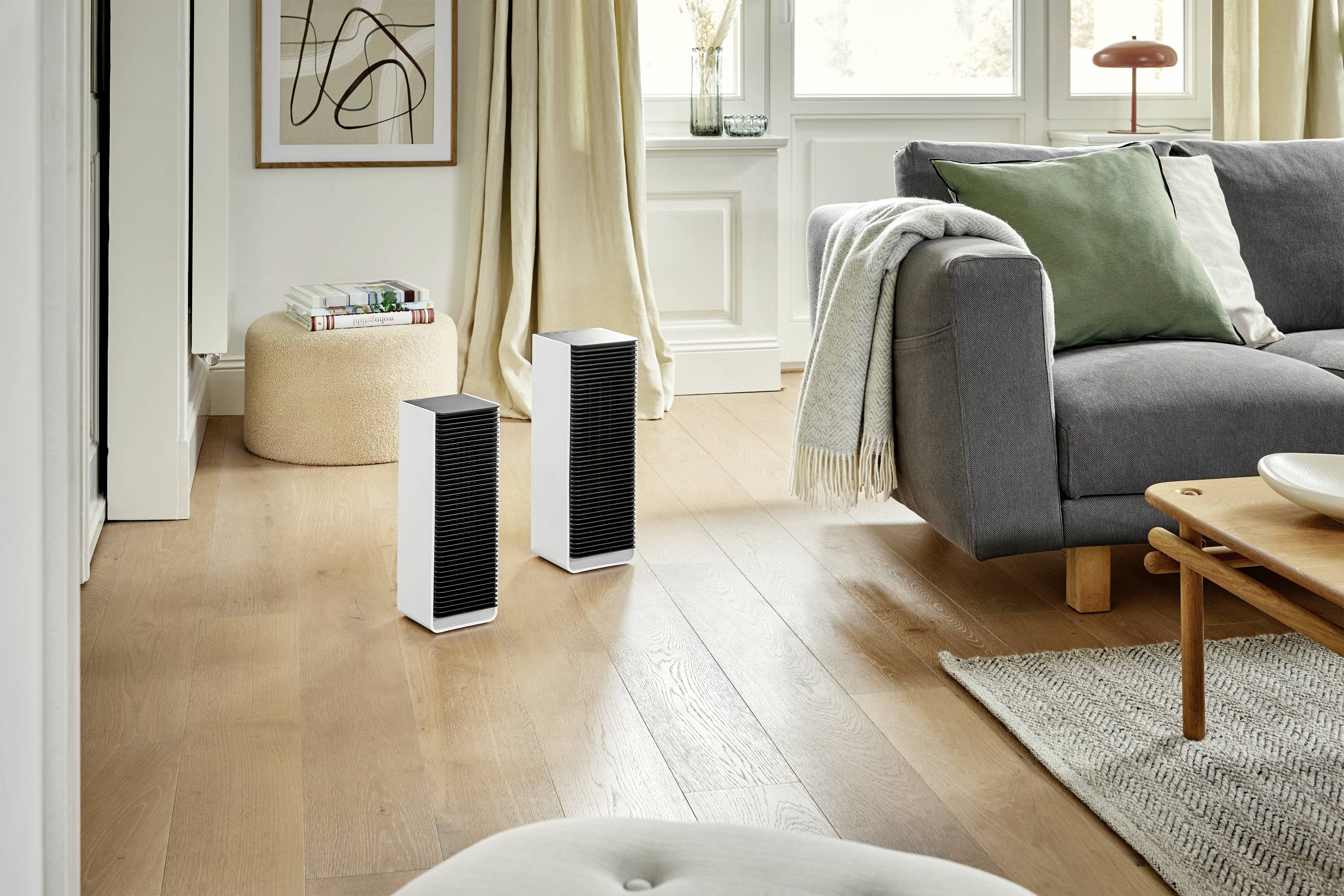 Two black speakers are standing on a wooden floor in a living room with a grey sofa and a green cushion.
