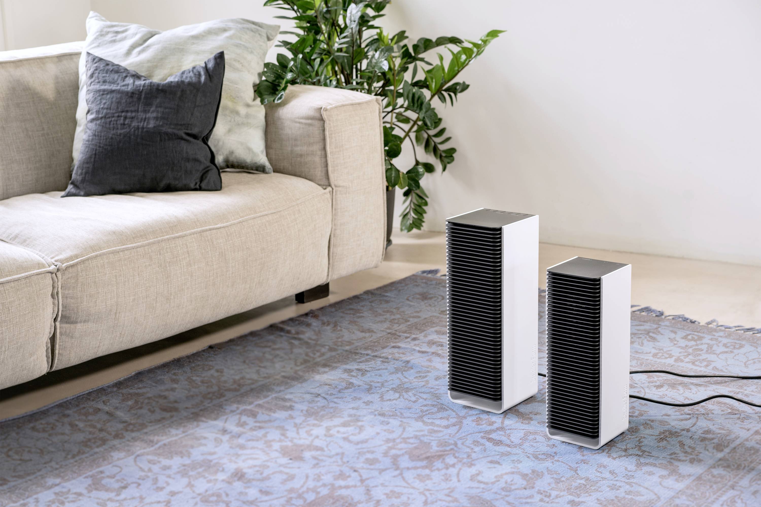 Two white air purifiers are standing side by side on a blue carpet in front of a beige sofa, next to a green plant.