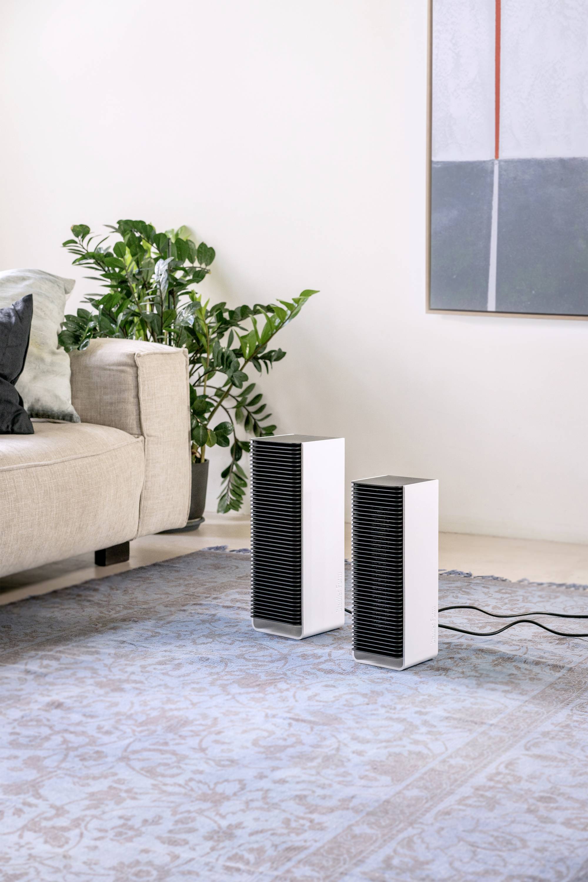 Two modern air purifiers stand on a carpet in a stylishly furnished living room with a sofa, plants, and a painting.