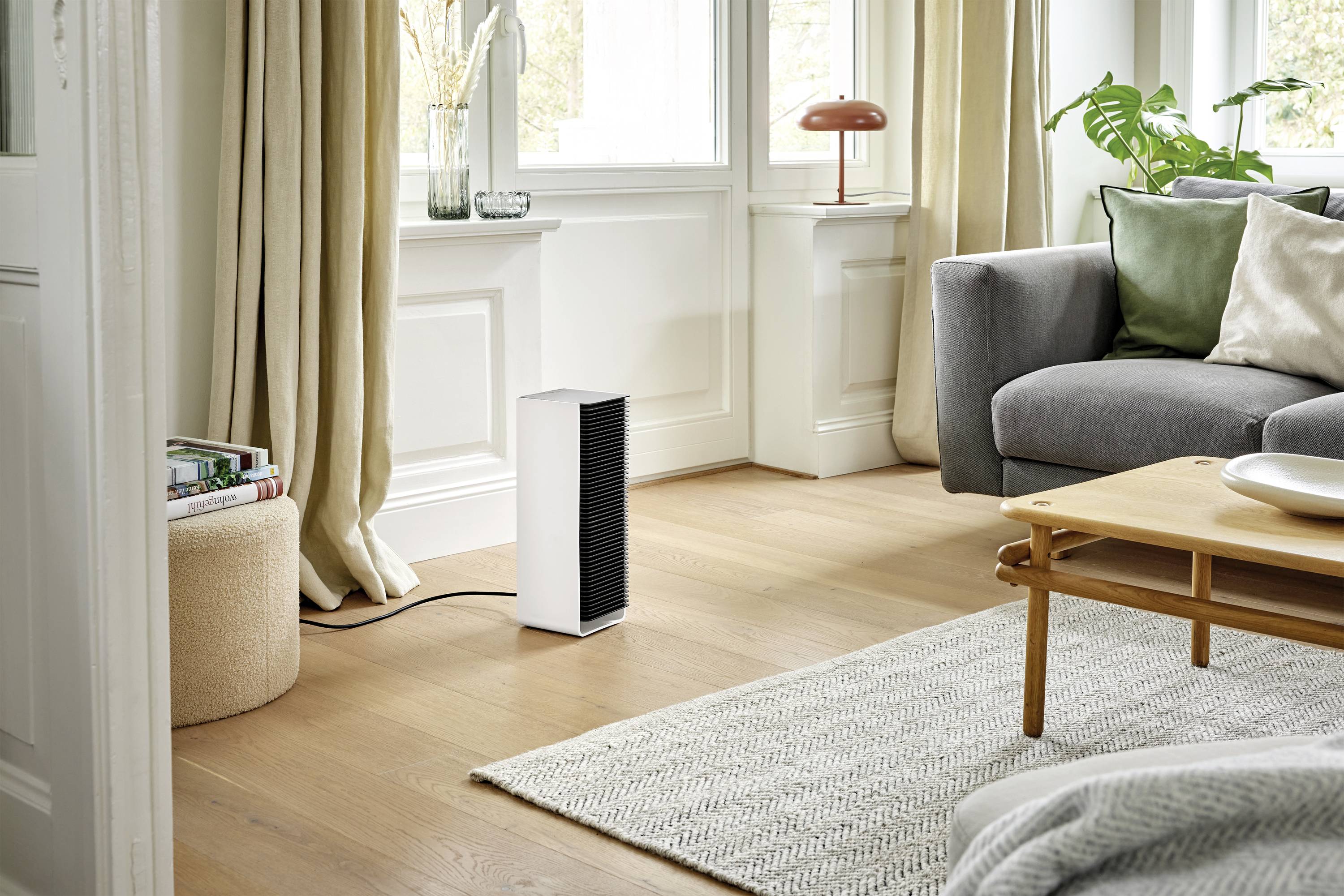A modern living room with an air purifier on the wooden floor, a grey sofa, a round table and large windows in the background.