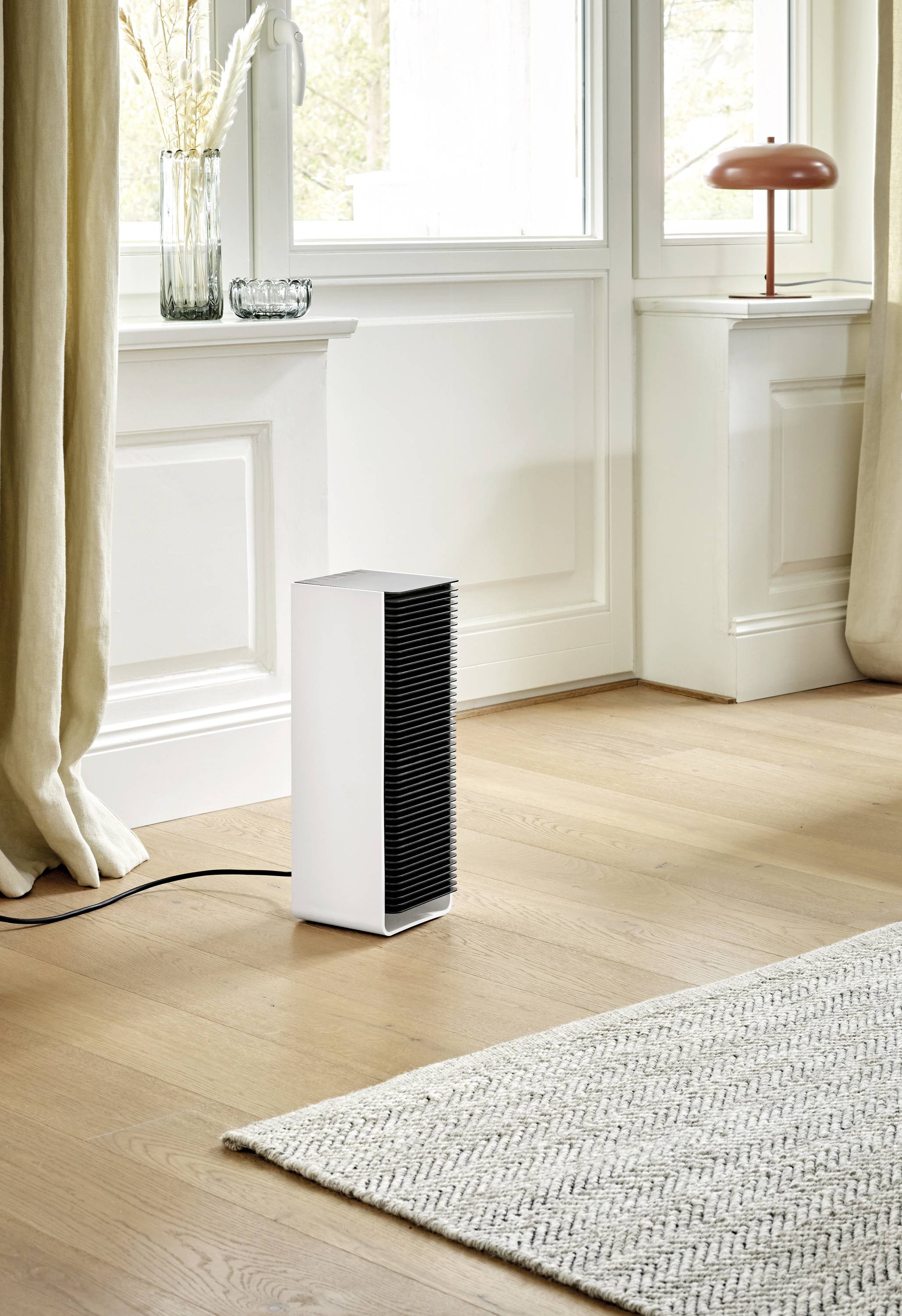 A white, rectangular fan heater stands on a wooden floor next to a carpet in a light-filled room with large windows.