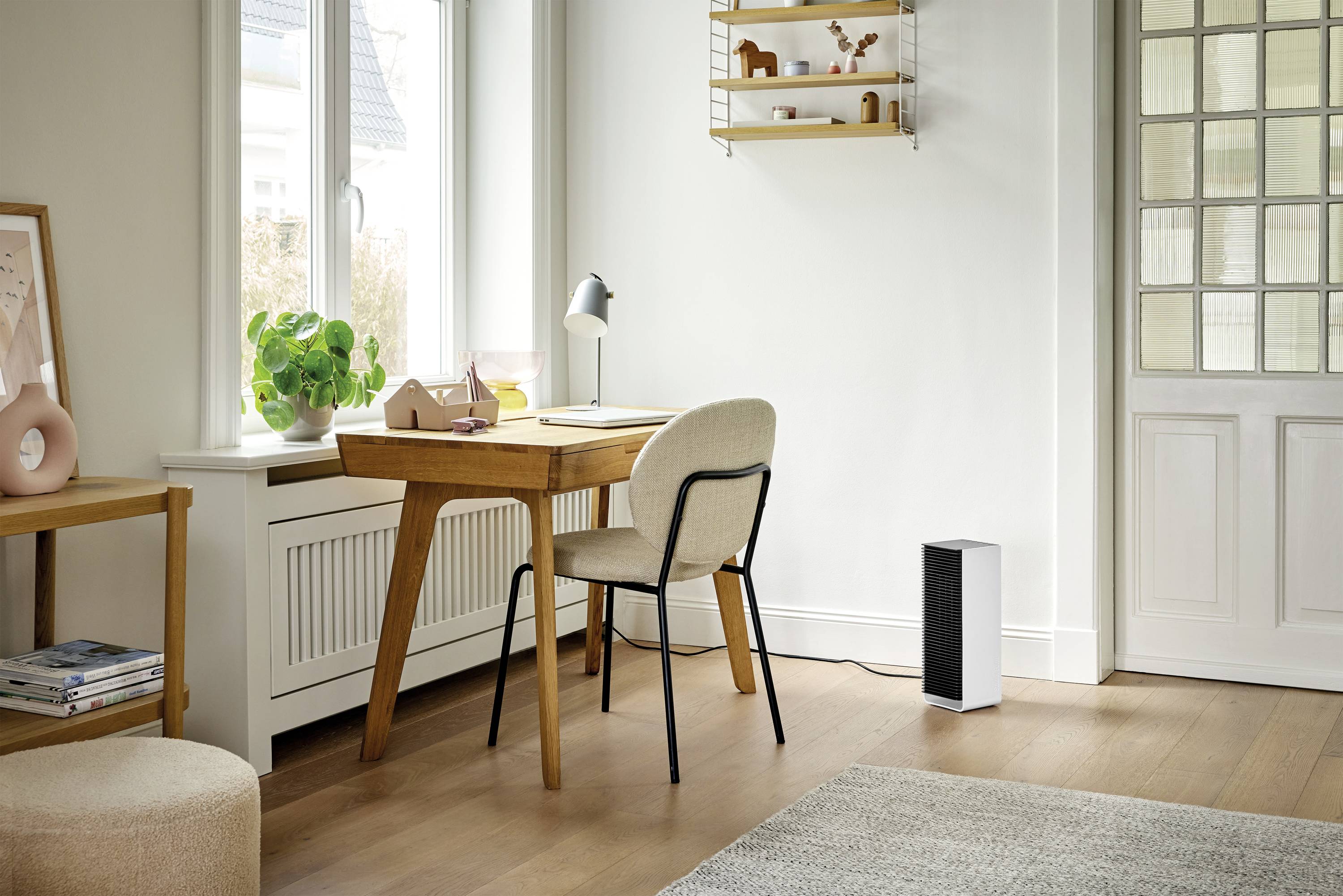 A bright, minimalist home office with a wooden desk, beige chair, plants, desk lamp and air purifier in front of a window.