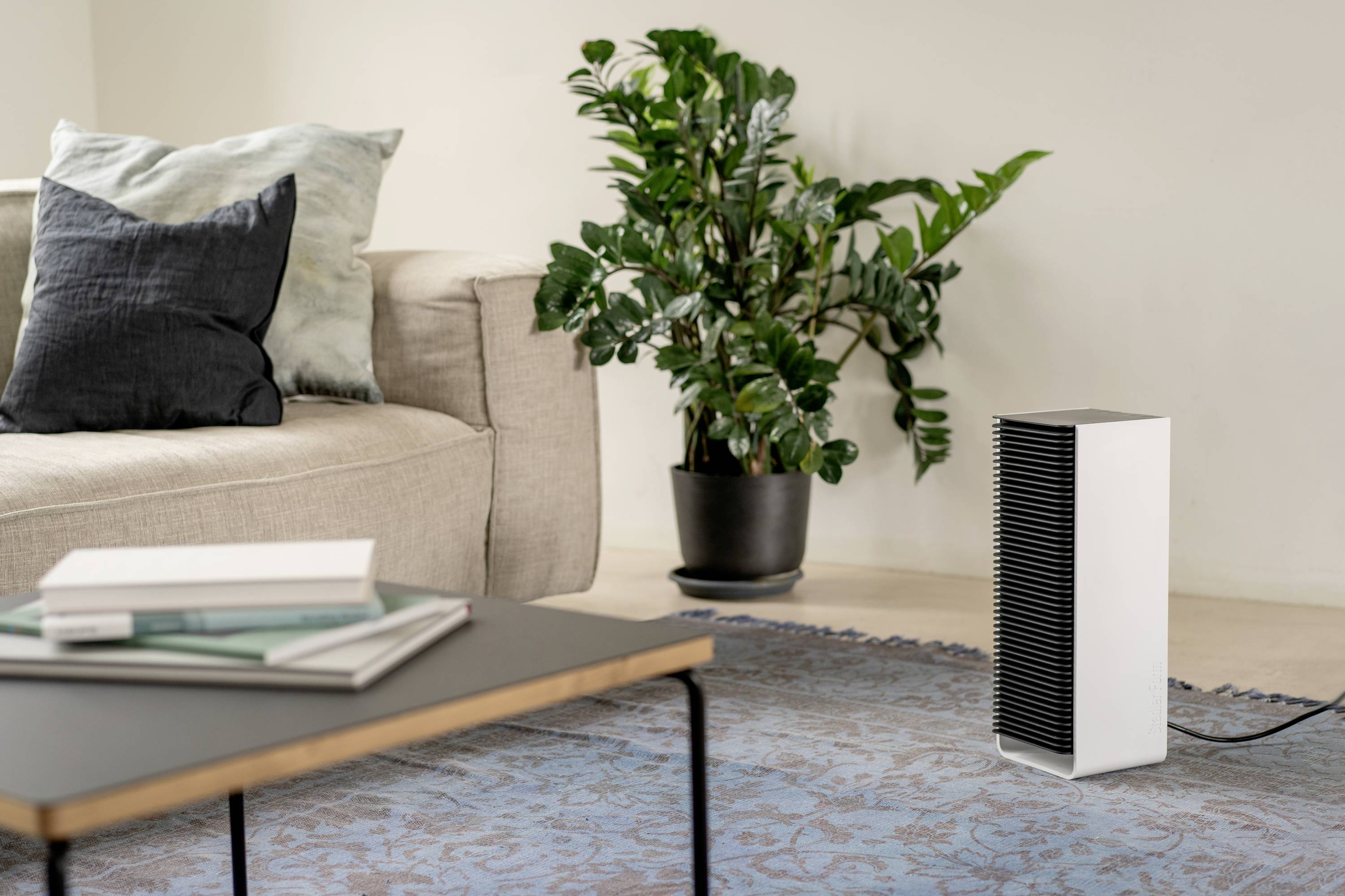 A modern air purifier stands on a carpet-covered floor next to a sofa and a plant in a stylishly decorated living room.