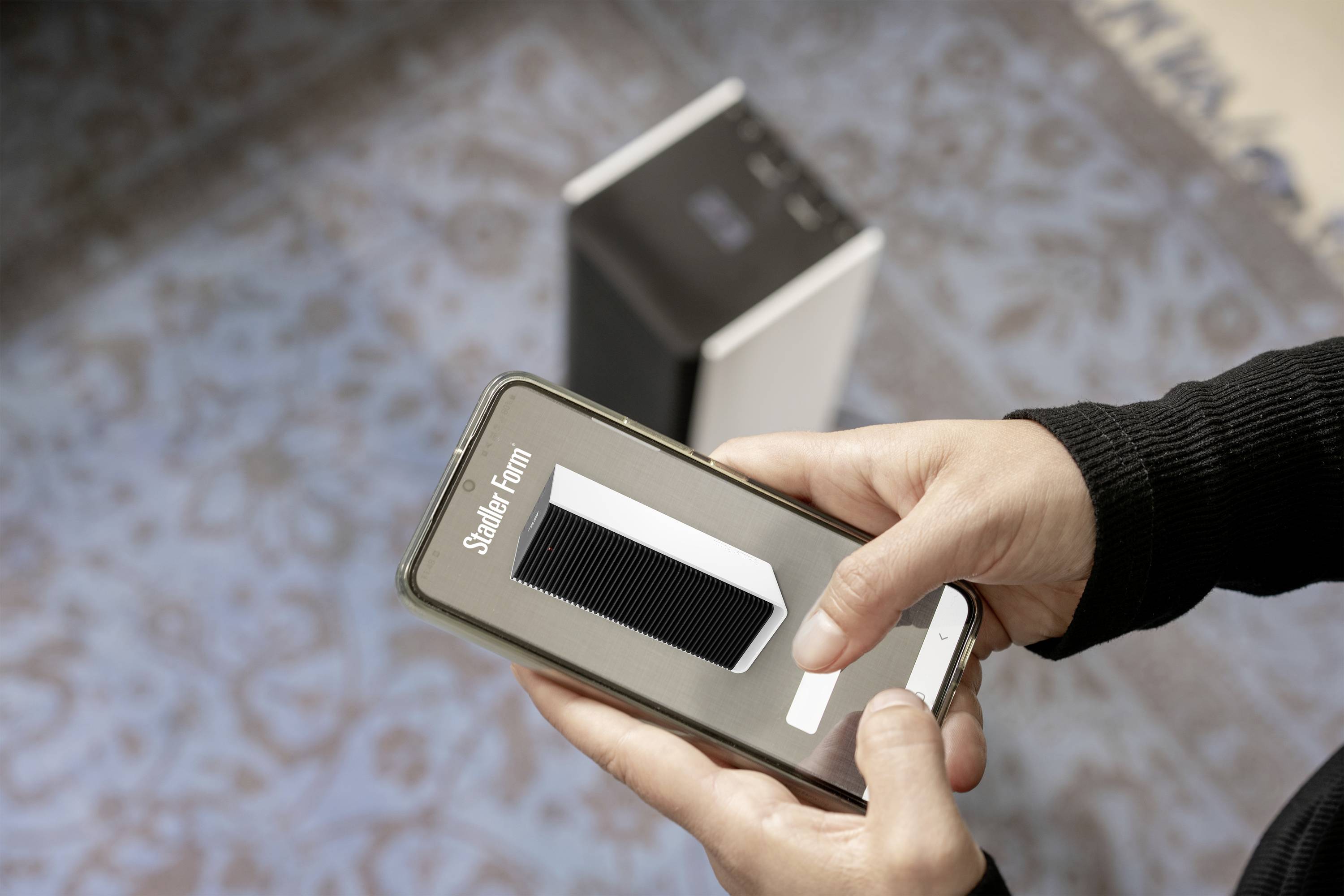 A person is holding a smartphone displaying an image of a speaker while standing next to a similar speaker.
