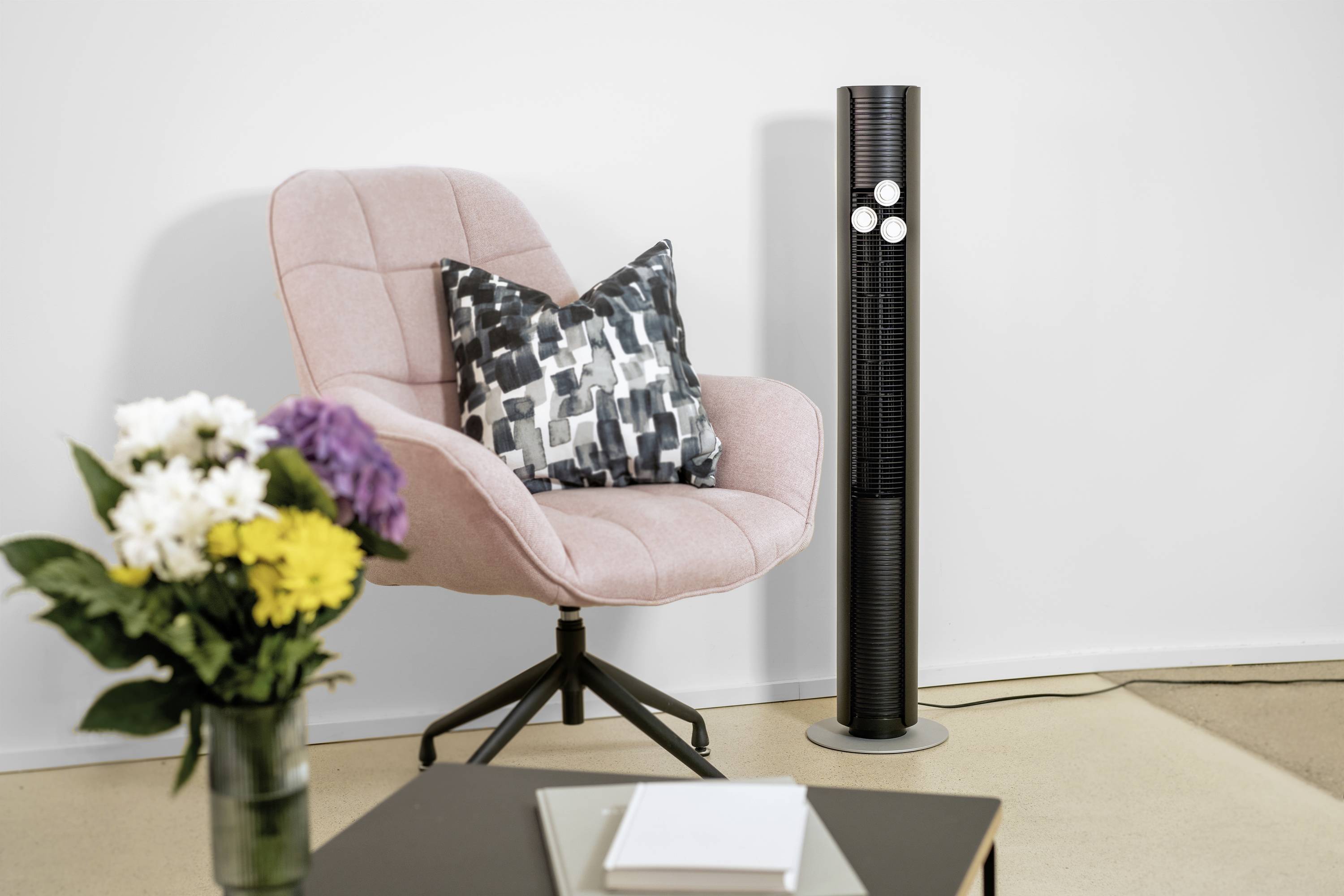 A pink armchair with a patterned cushion beside a black tower fan. In the foreground, a table with stacks of books and flowers in a vase.