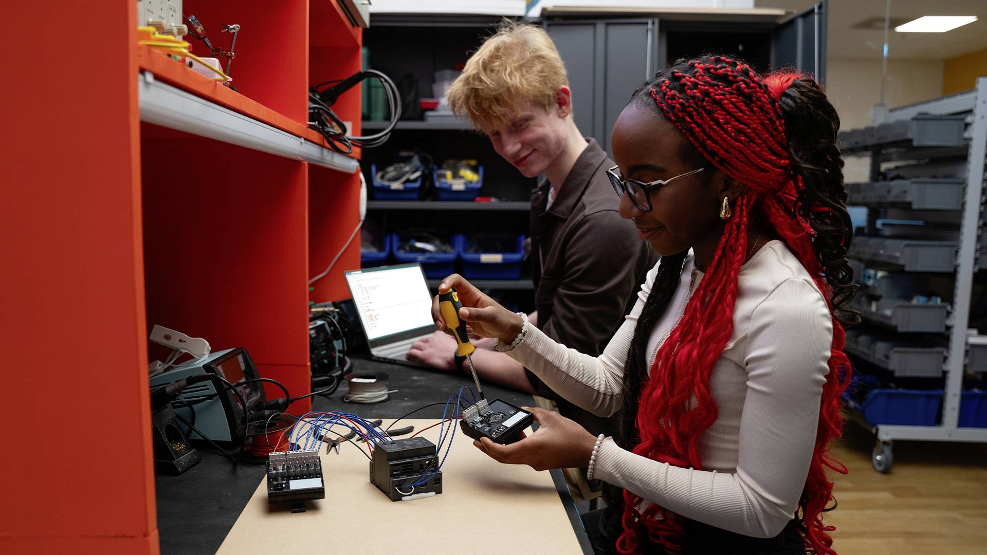 Two people are working in a technical laboratory with electronic equipment. A woman is holding a tool, while a man sits at the laptop.
