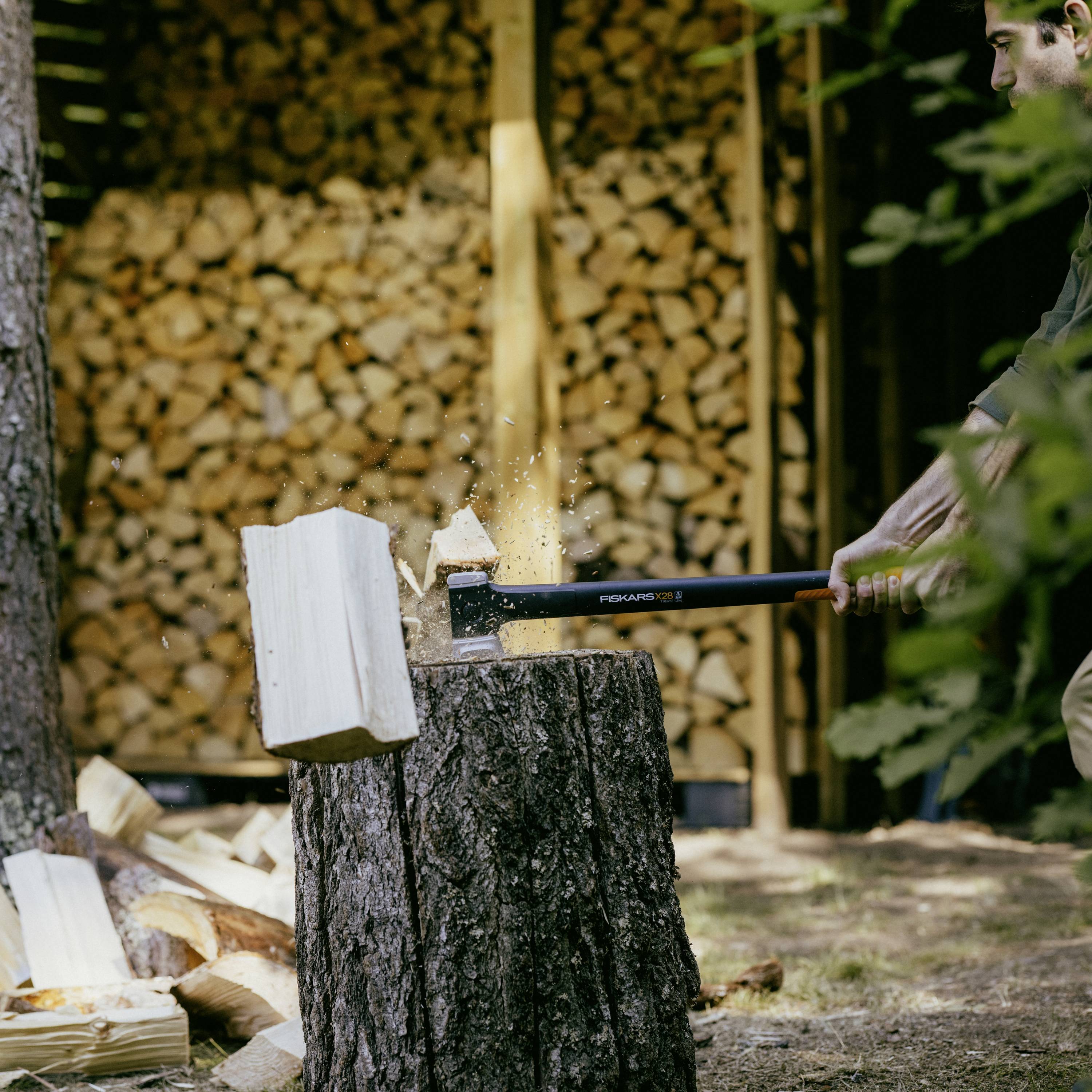 A person is splitting a log with an axe on a tree stump. Stacked piles of wood can be seen in the background.