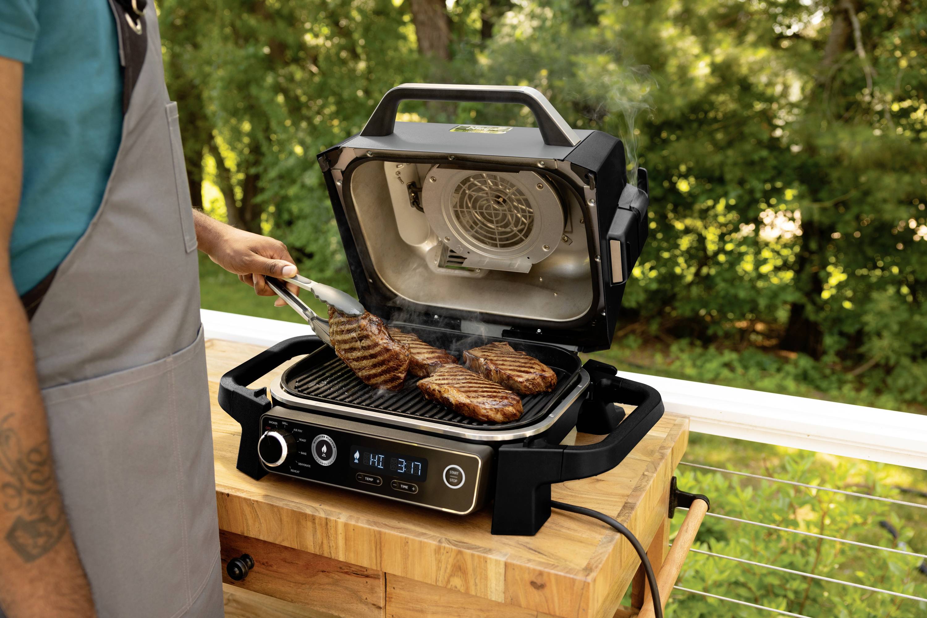 A person is grilling steaks on an electric barbecue outdoors. The grill displays a temperature gauge, with plenty of greenery visible in the background.