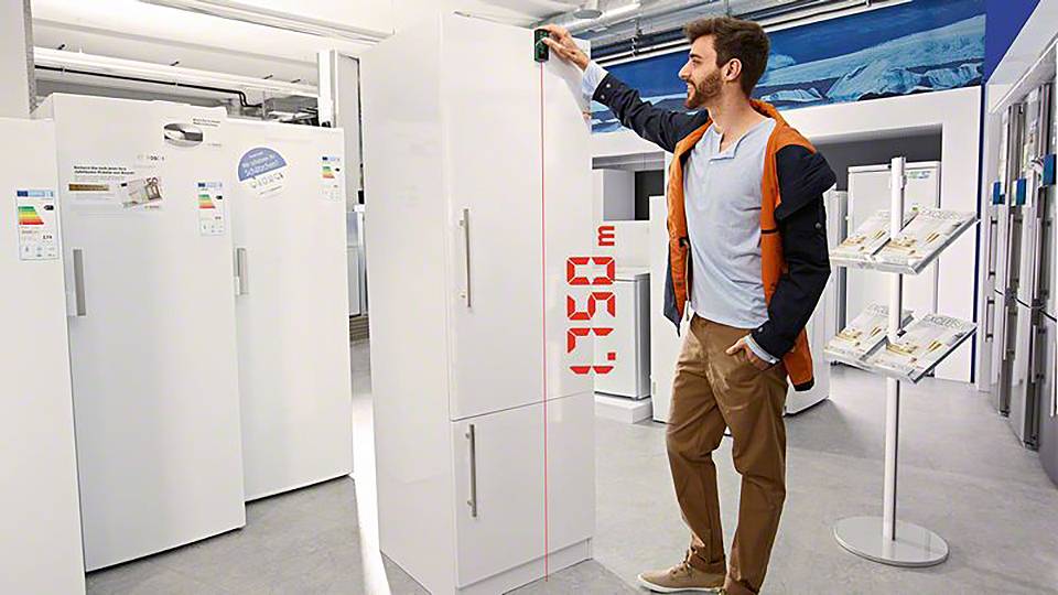 A person is measuring the height of a white fridge with a laser device in an electrical appliance shop.