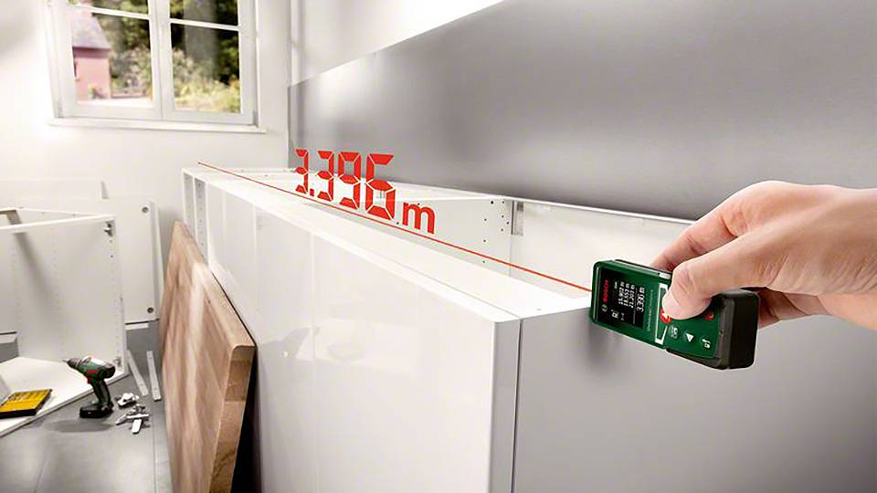 A hand is holding a laser distance meter displaying a distance of 3.395 m on a kitchen worktop. A window is visible in the background.