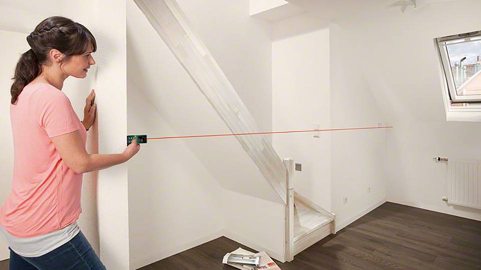 A woman is measuring the distance in an attic room using a laser distance metre. An angled roof window is visible.