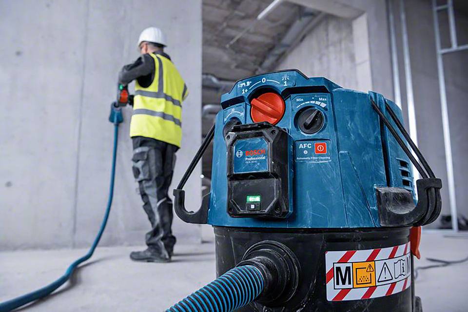 A construction worker in protective clothing is using an industrial vacuum cleaner on a building site. The vacuum cleaner is in the foreground.