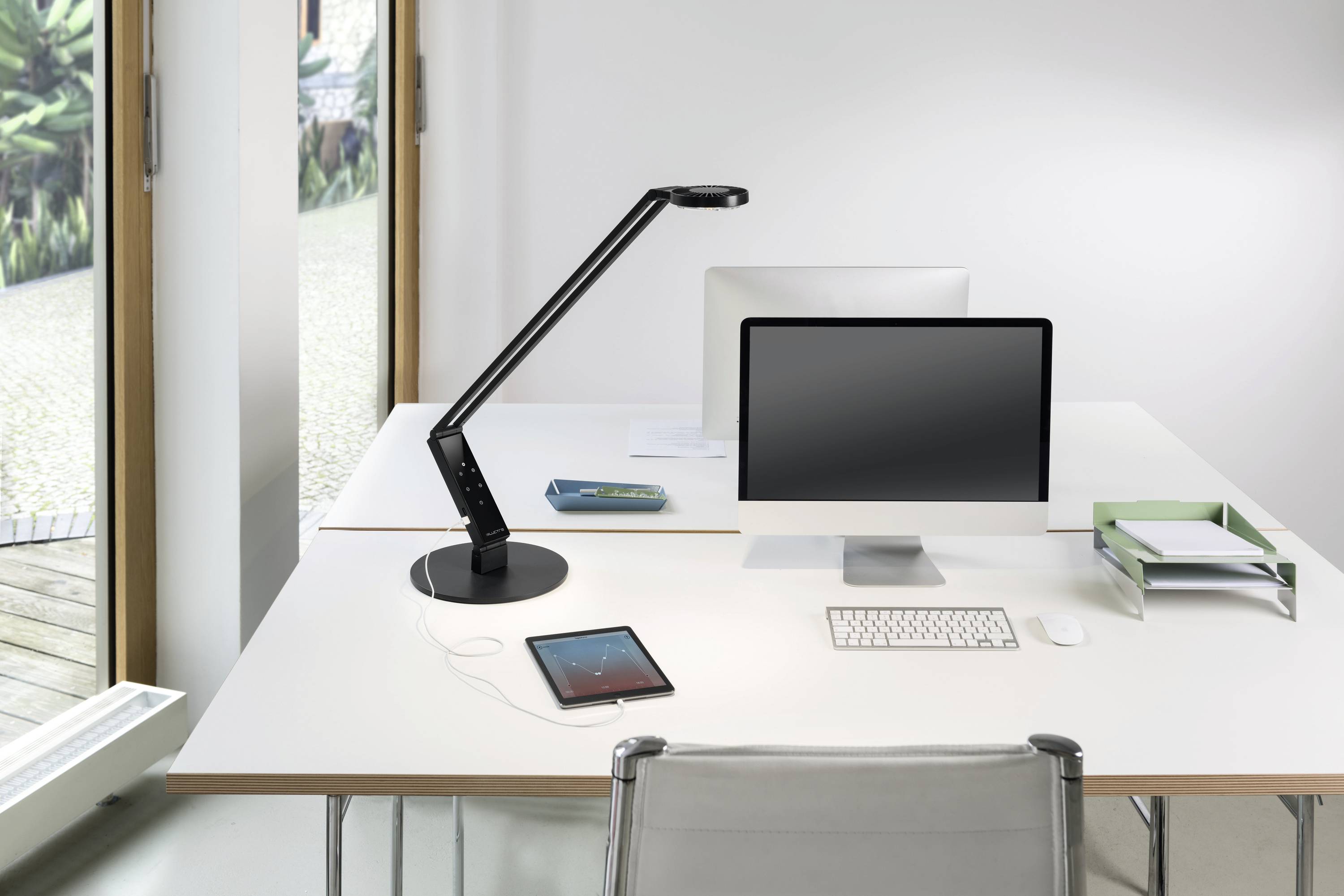 A tidy desk with a monitor, a desk lamp, a tablet, a book and a paper tray beside a window.