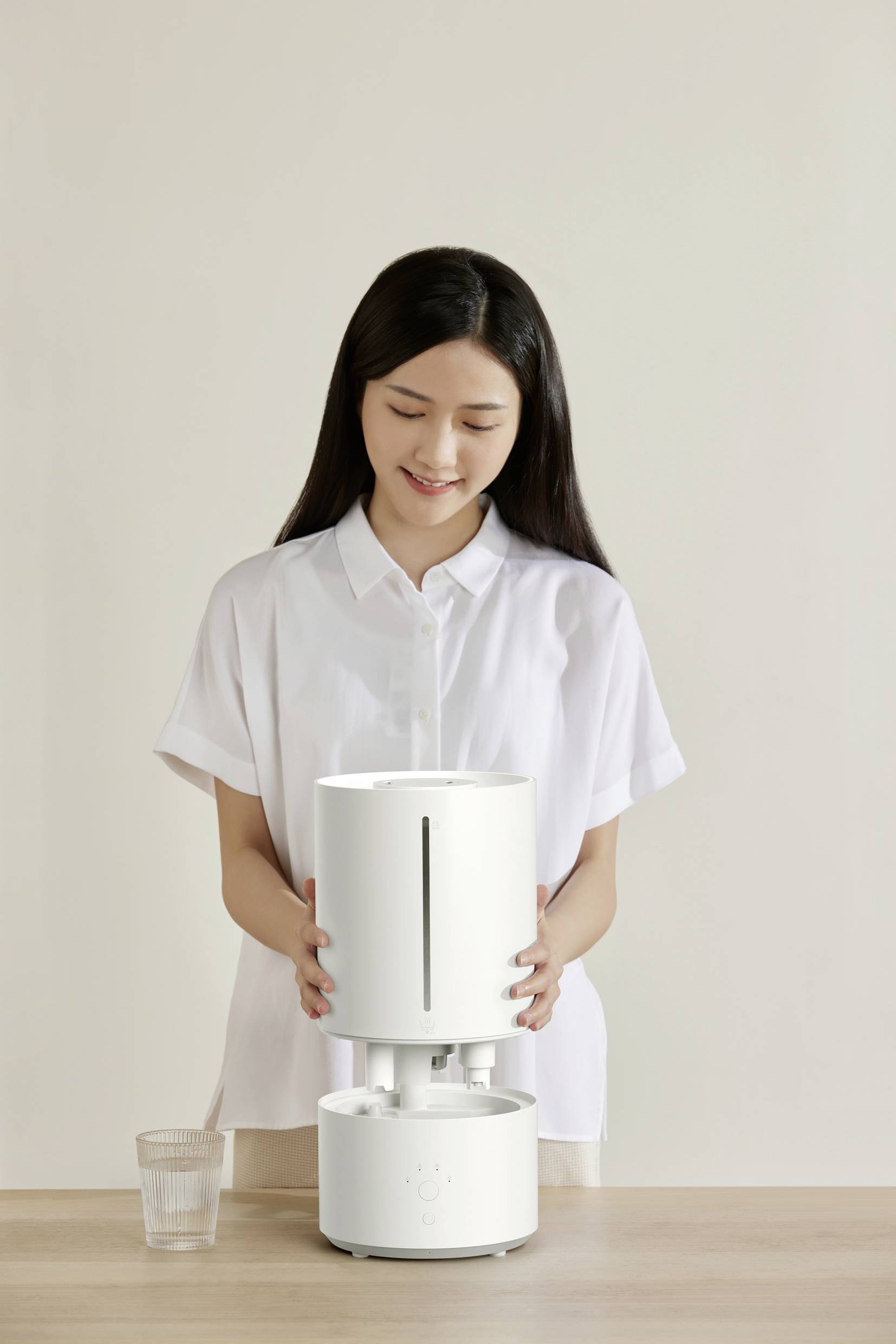 A woman in a white shirt is smiling while operating a humidifier placed on a table. A glass of water sits beside it.