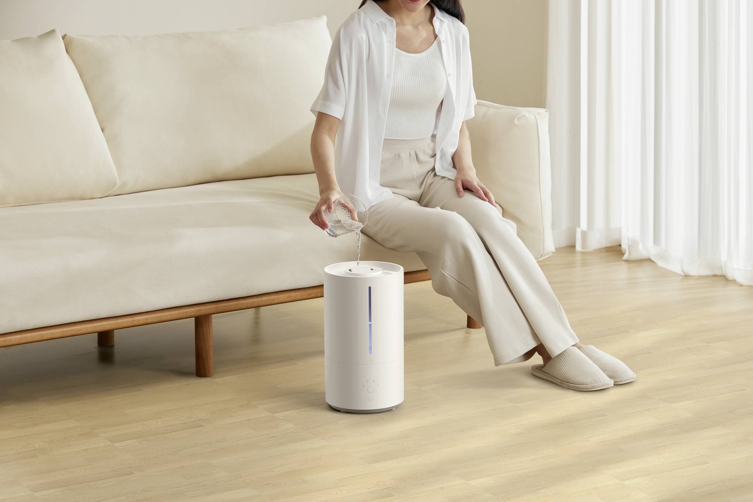 A woman is sitting on a sofa and pouring water into a humidifier that is standing on the wooden floor. She is wearing light-coloured clothing.