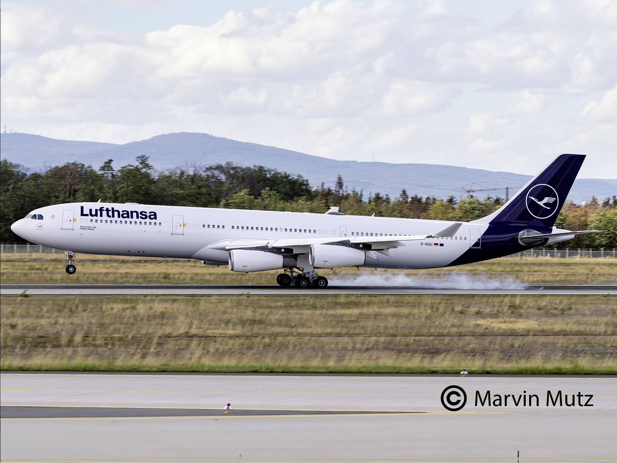 A Lufthansa aircraft, an Airbus A340, is landing on a runway with smoke from its tyres. Mountains are visible in the background.
