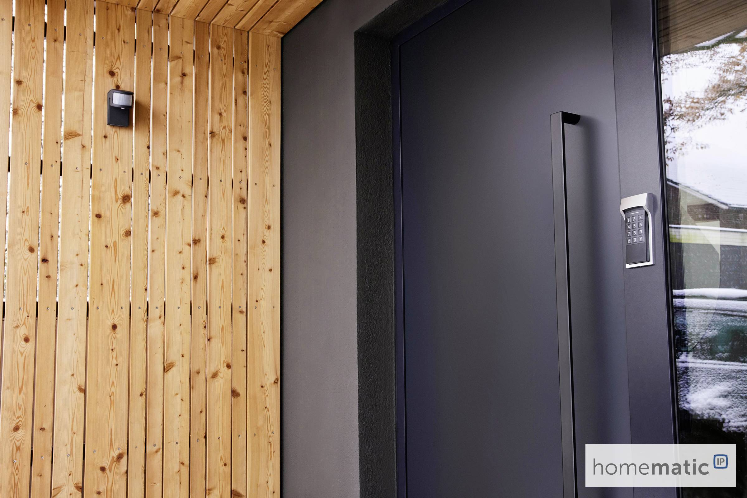 Image shows a modern front door with a code lock. To the left is a wooden panelling. The 'homematic' logo is located in the bottom right.