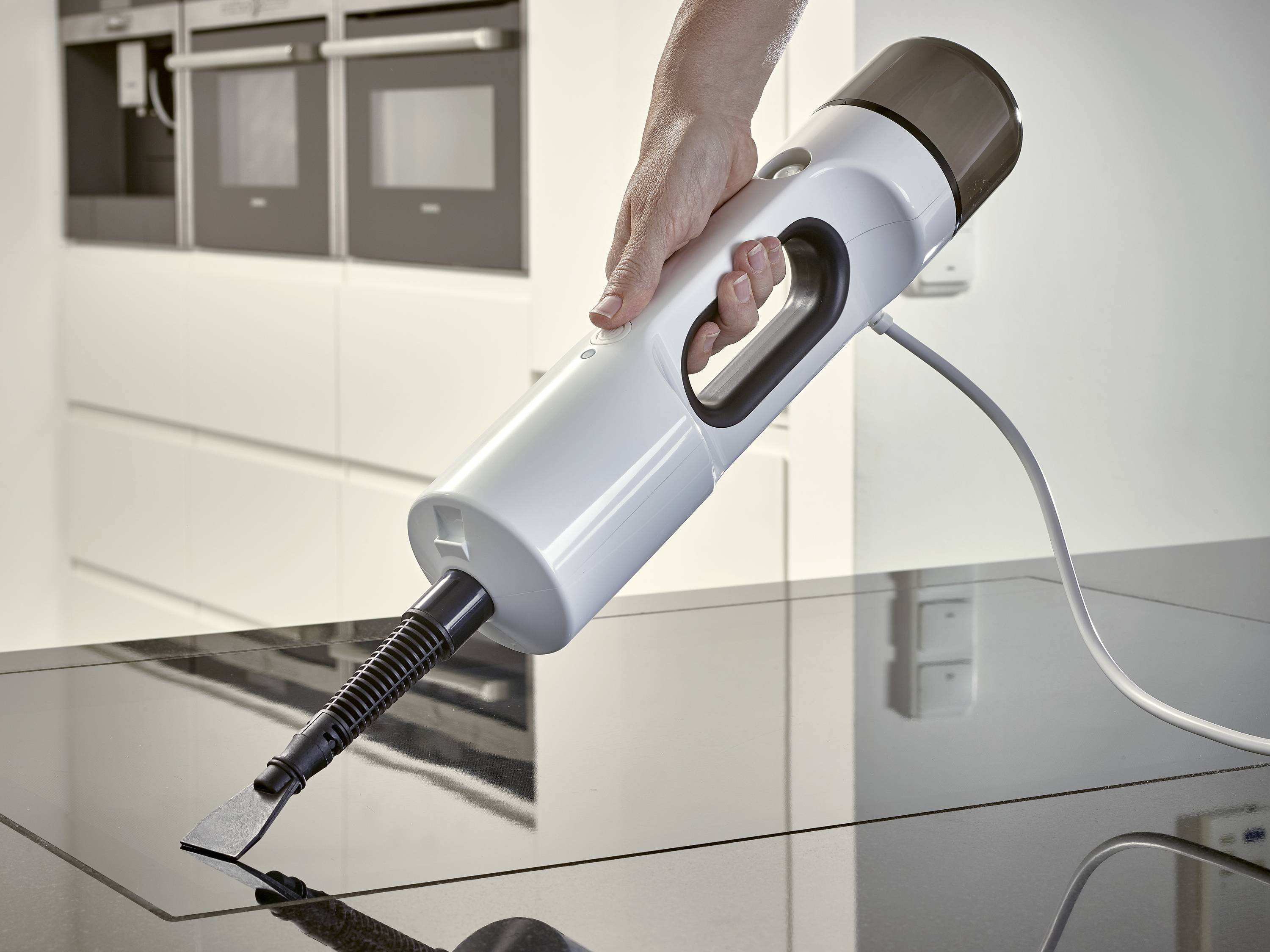 A hand uses a steam cleaner to clean a smooth, dark kitchen surface. Kitchen cupboards are visible in the background.