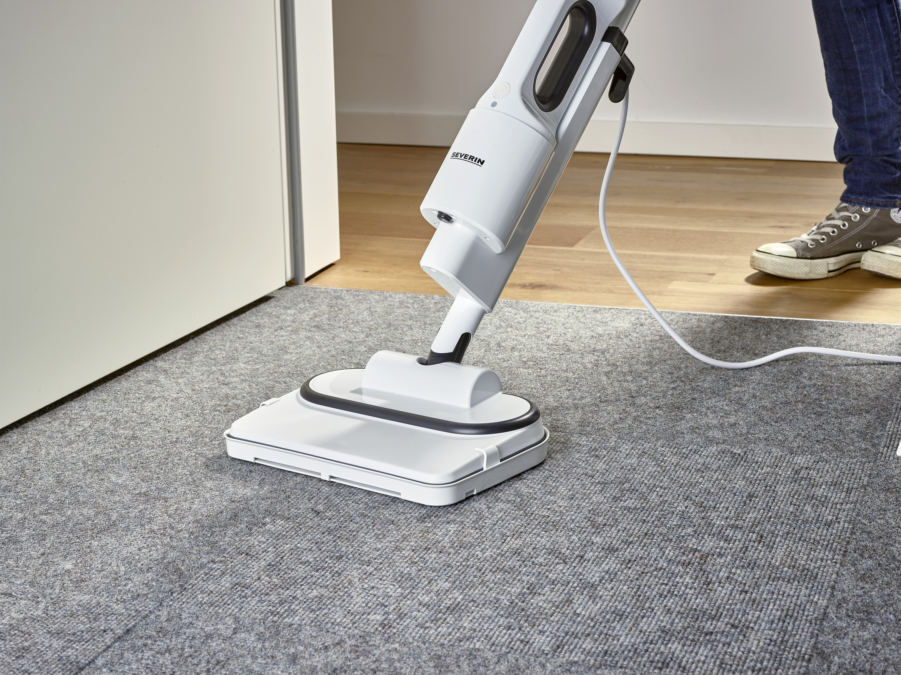 A person is cleaning a grey carpet with a white, corded vacuum cleaner in a room with wooden flooring.