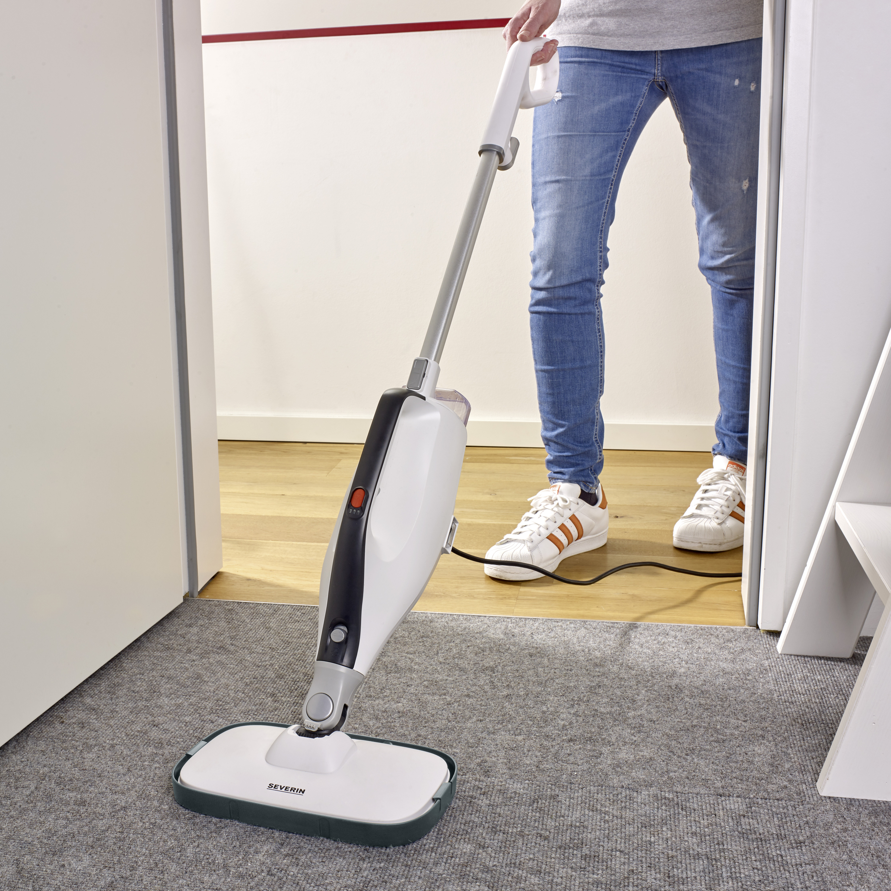 A person is using a steam cleaner on a carpet. The floor cleaner has a modern design and is in operation, while the person is wearing jeans and trainers.