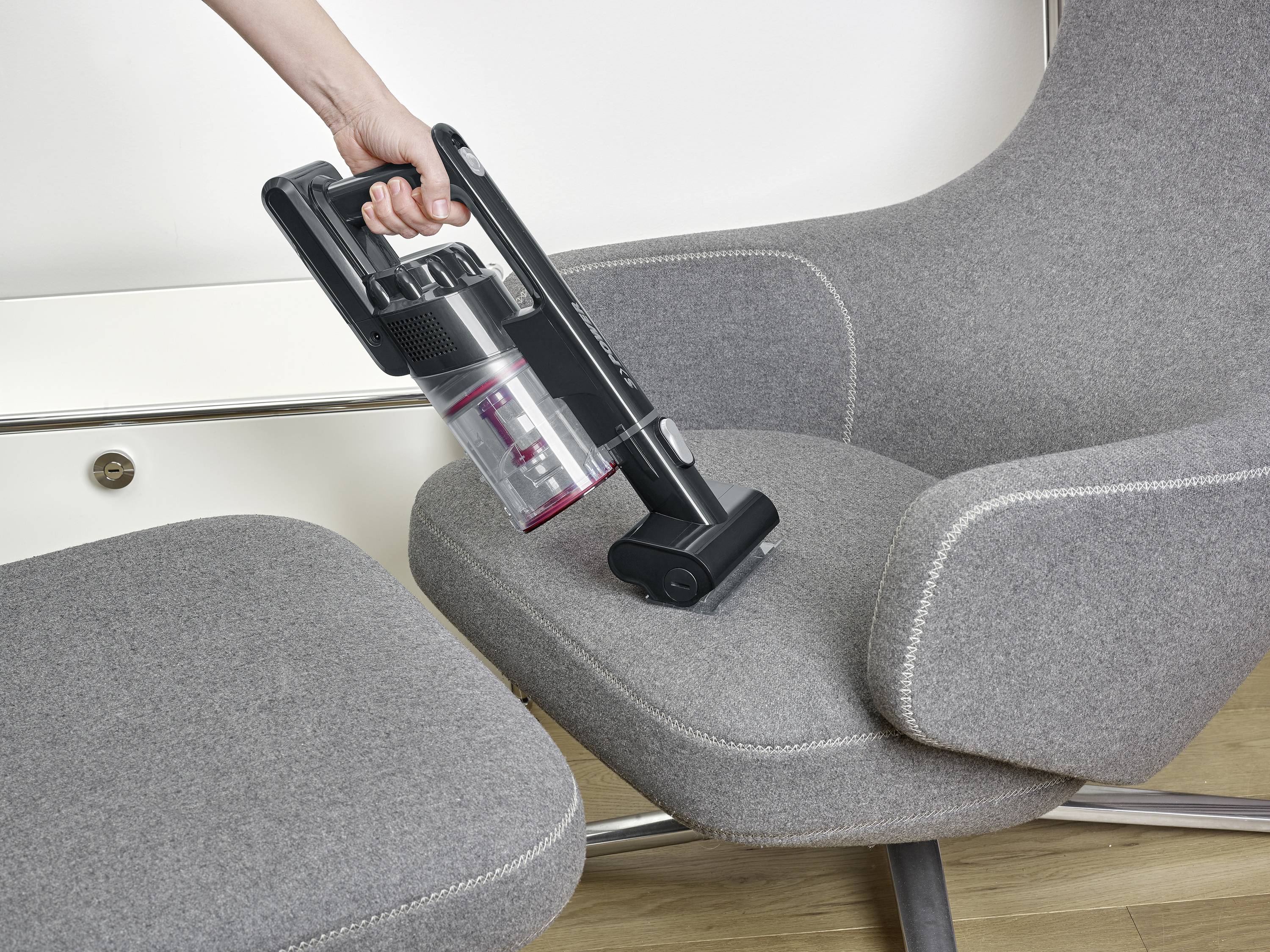 A person is cleaning a grey chair with a cordless handheld vacuum cleaner. The chair has a smooth, minimalist design.