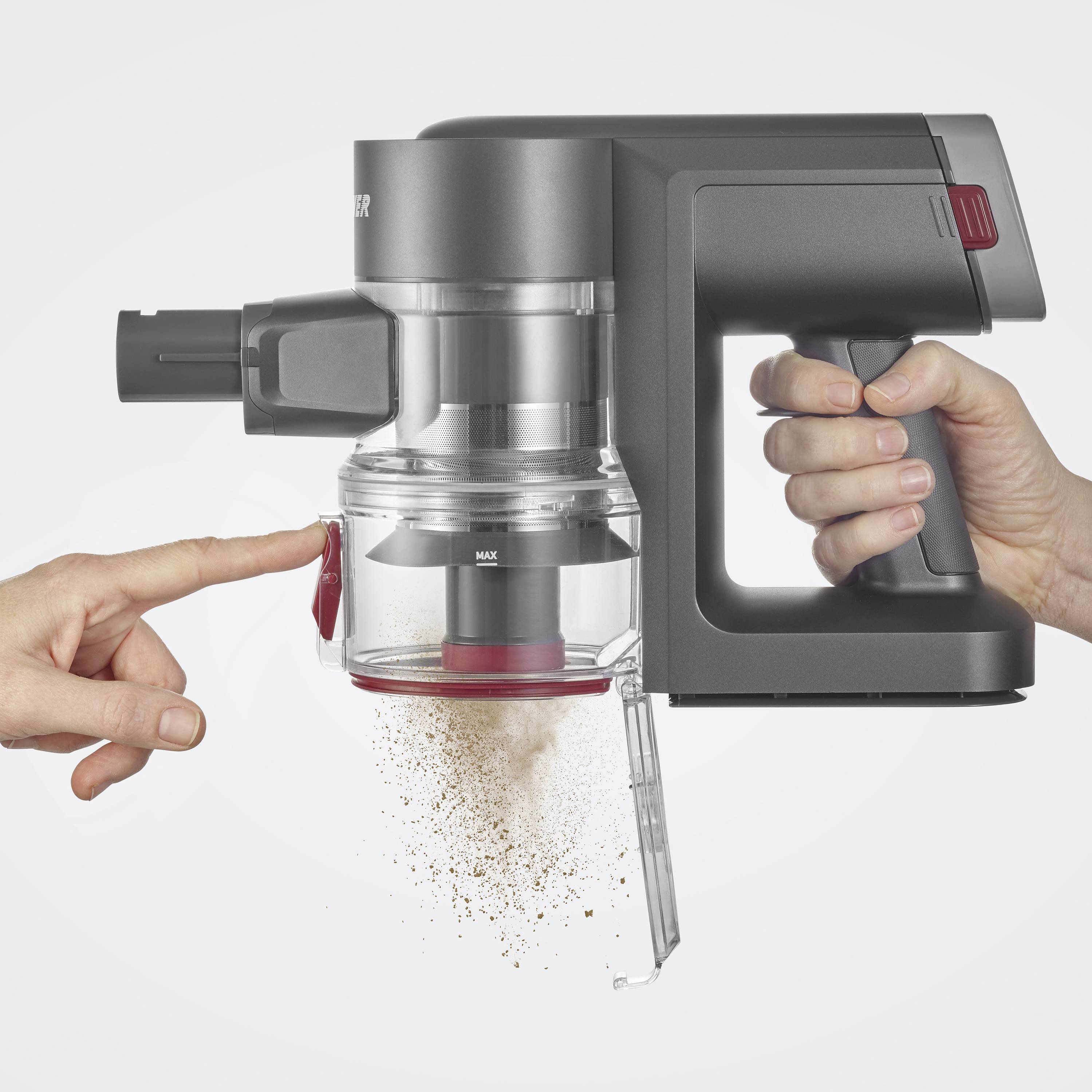 A vacuum cleaner is being held, with dirt falling out of the open container. A hand is pointing to the emptying mechanism.