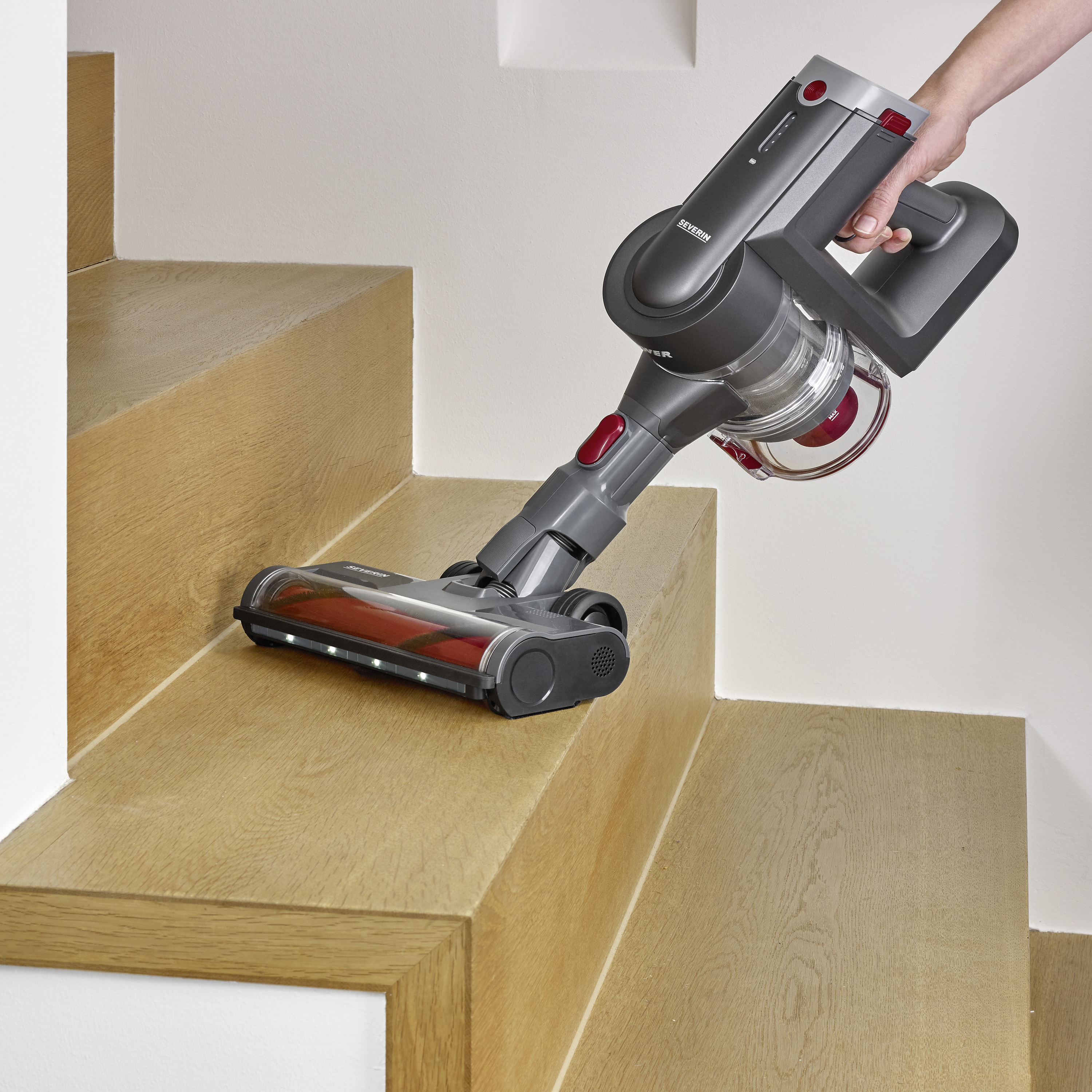 A cordless vacuum cleaner is cleaning a wooden staircase. One hand is holding the vacuum cleaner handle, while the brush moves over the steps.