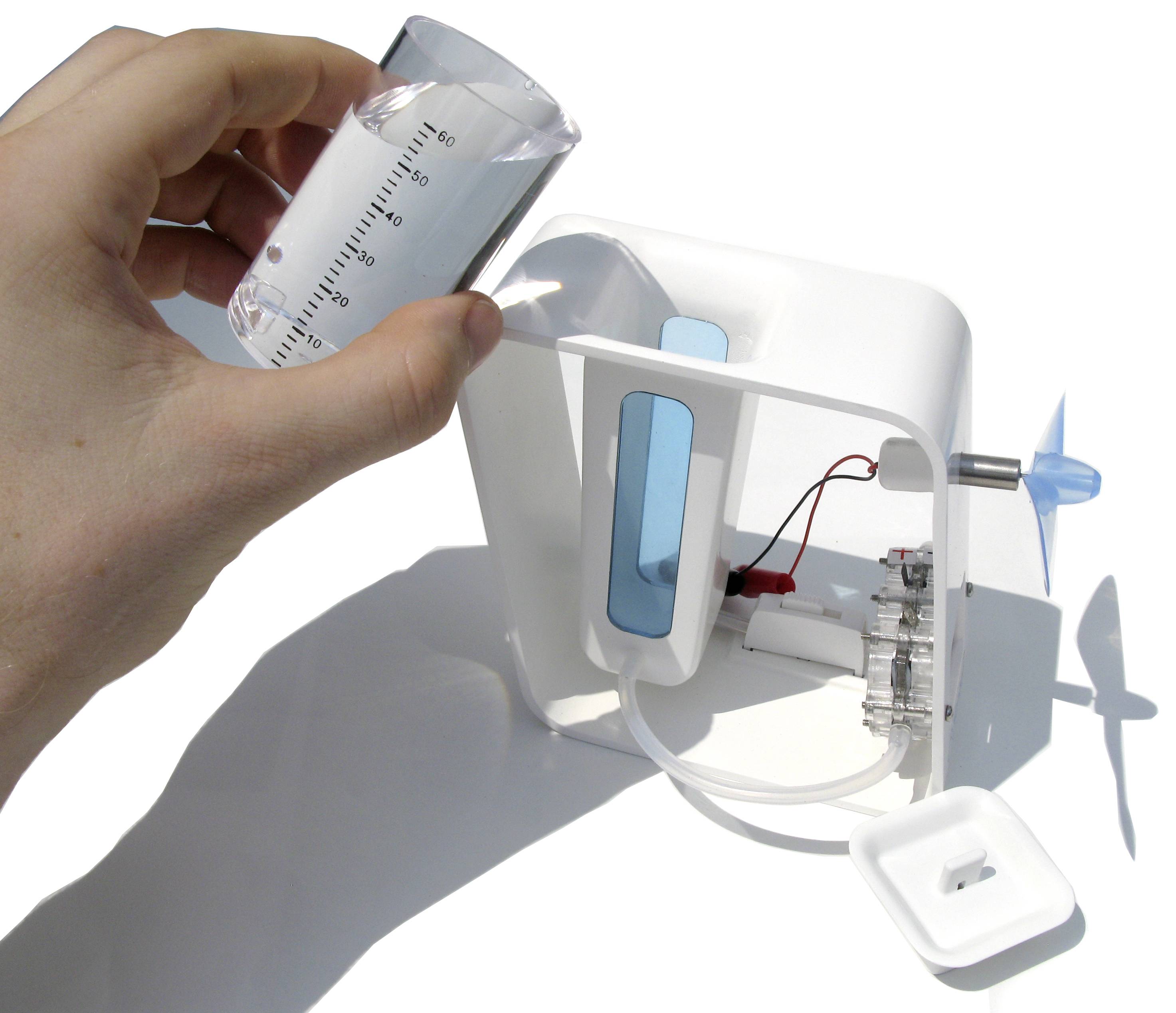 A hand holds a measuring cup over a mini wind turbine model with transparent blades, which is powered by water.