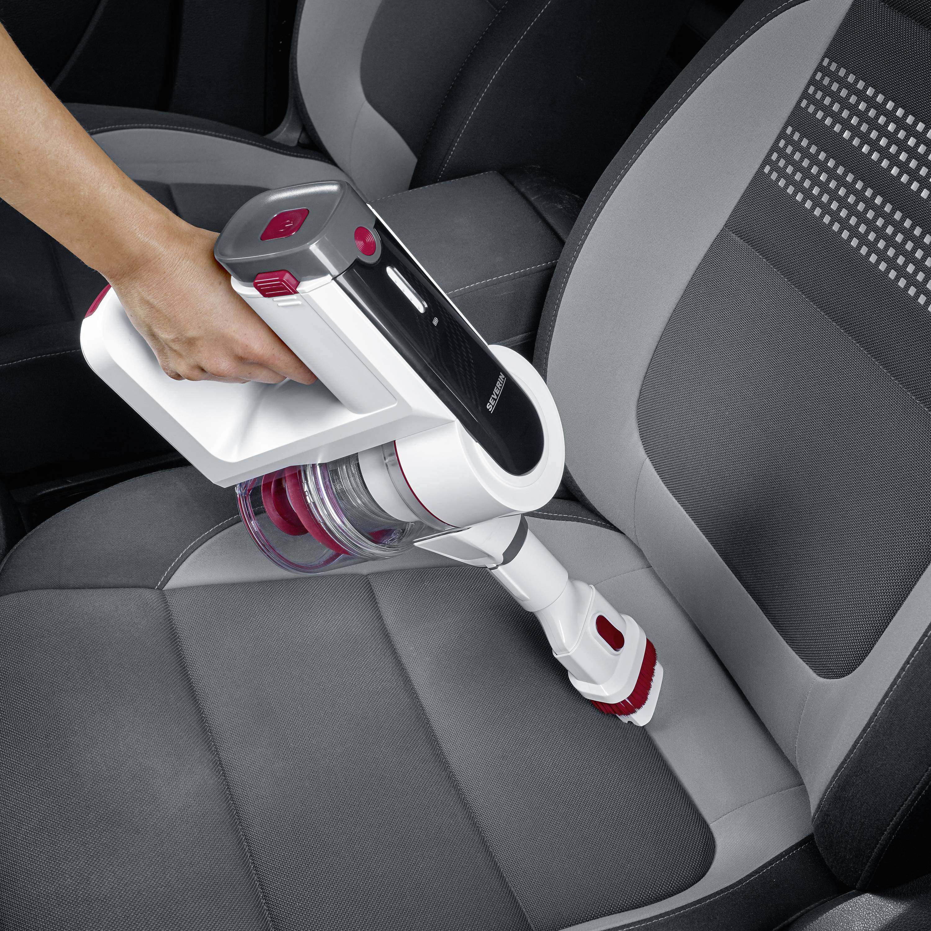 A person is cleaning the car seat with a cordless handheld vacuum cleaner. The seat is grey with black accents.