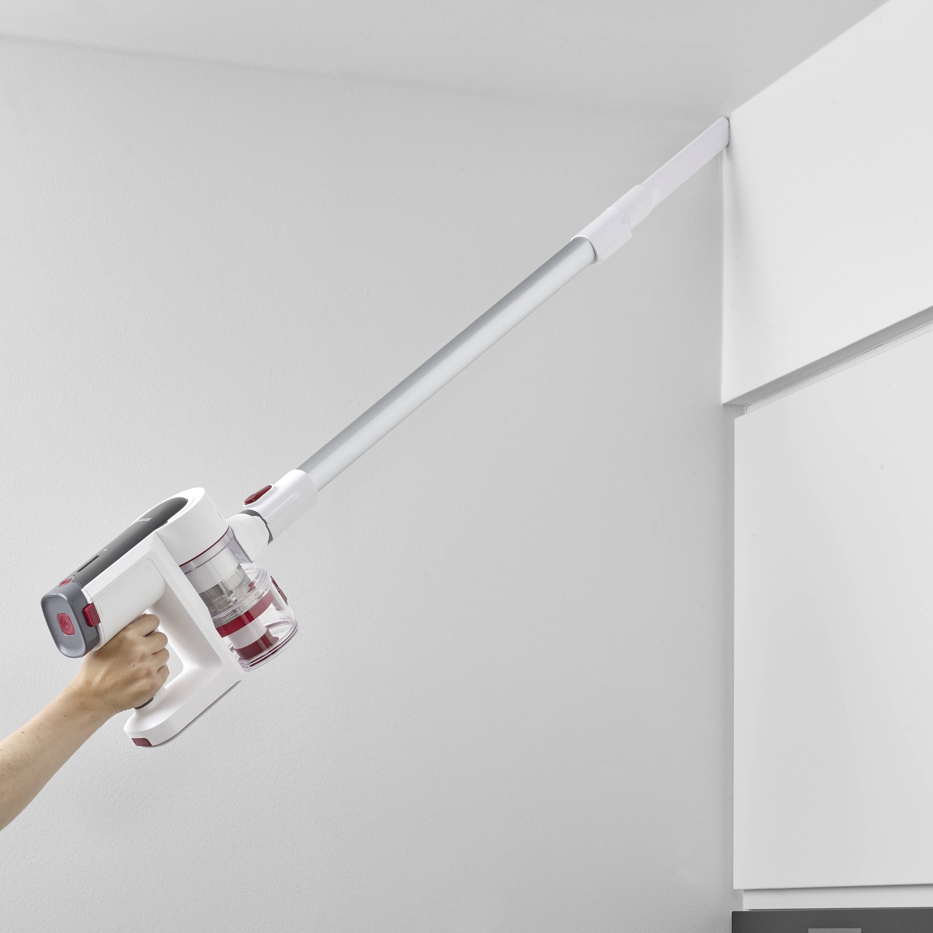A person is using a cordless vacuum cleaner with a long wand to clean a corner near the ceiling.