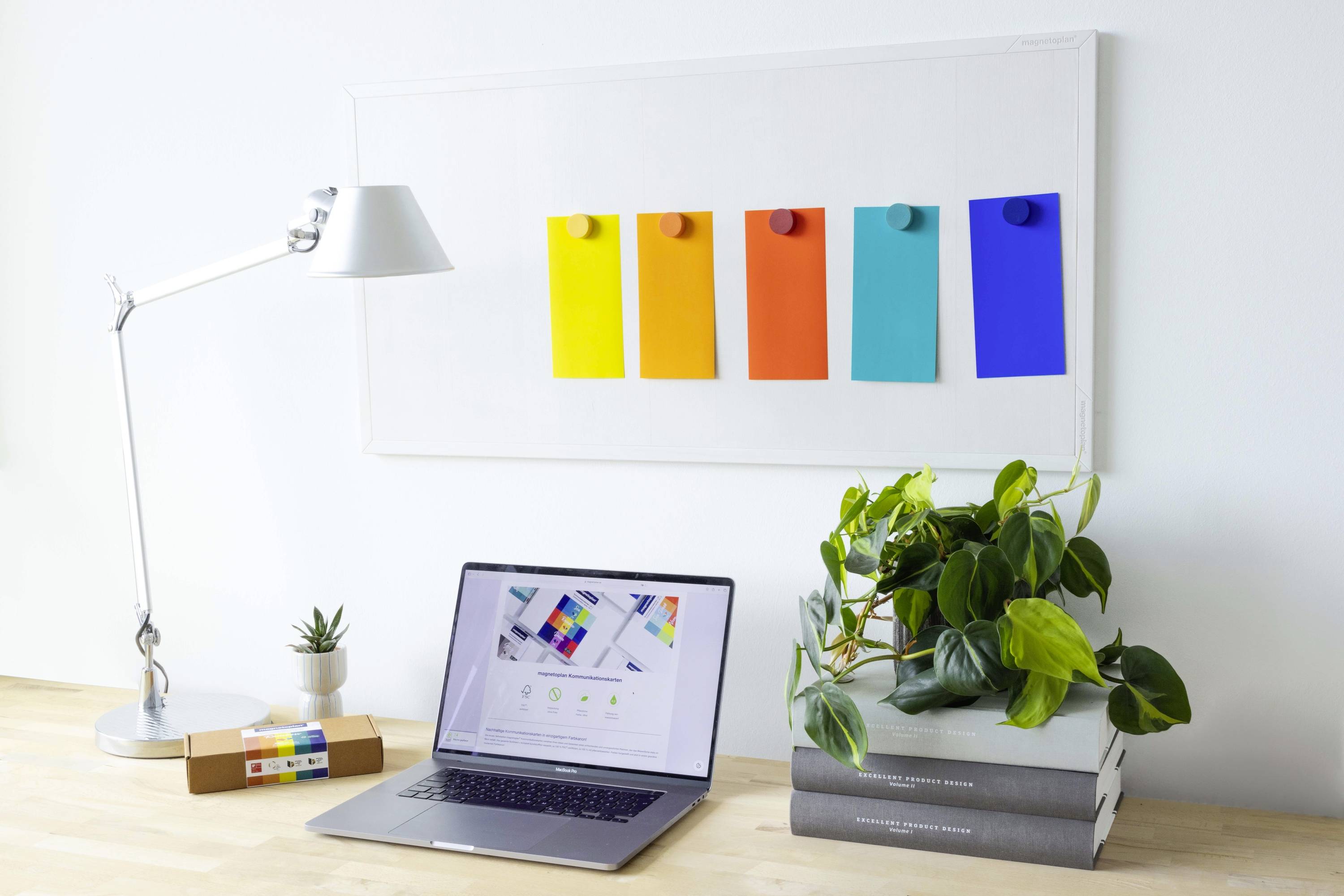 A desk with a laptop displays a website with colour cards. On the wall hang five colour swatches in yellow, orange, red, turquoise, and blue.