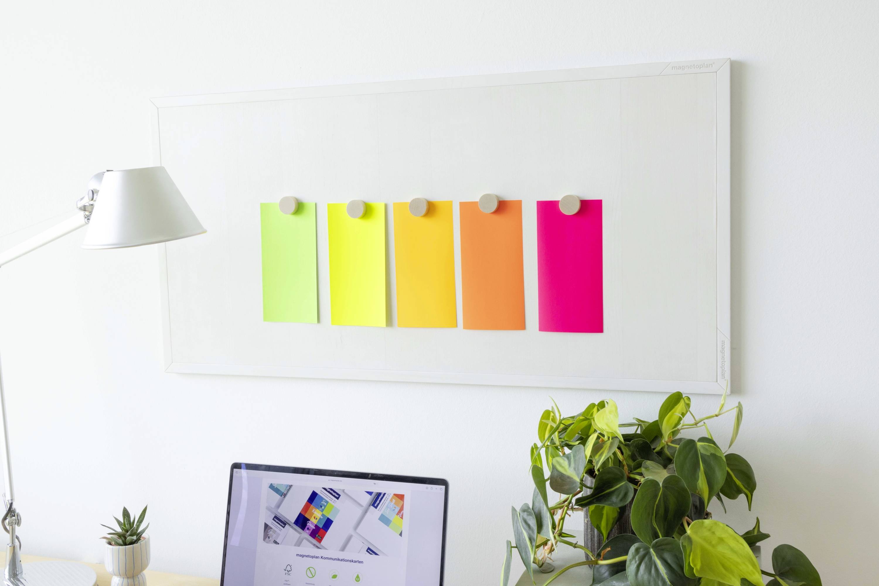 A desk with a lamp, laptop and plants in the foreground; in the background, a board hangs with colourful sticky notes.