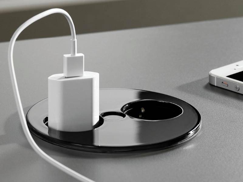 A smartphone charger plugged into a power outlet built into a desk, with a smartphone placed nearby.