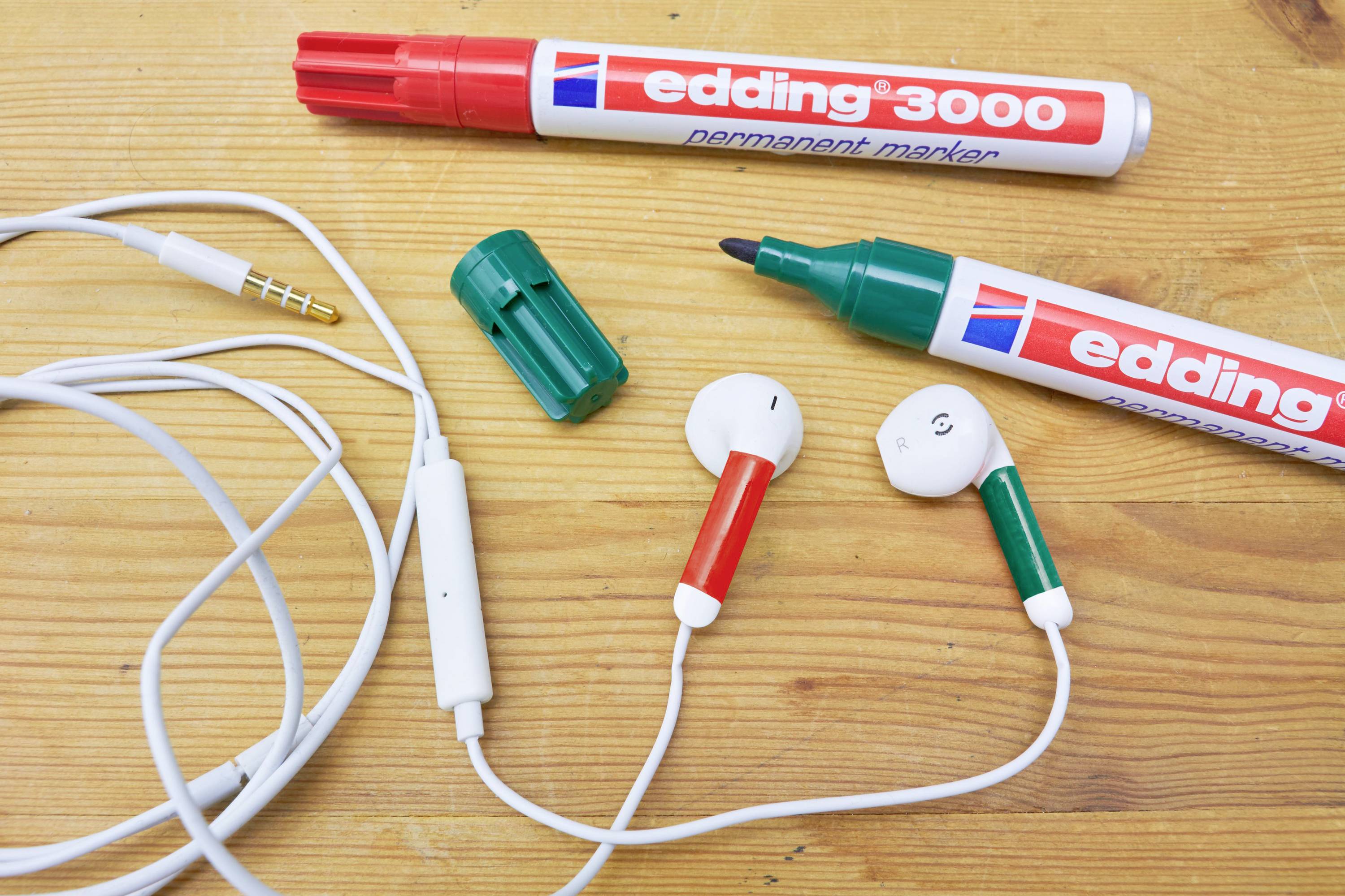 Two red Edding markers and a pair of white earphones with red and green elements are lying on a wooden table.