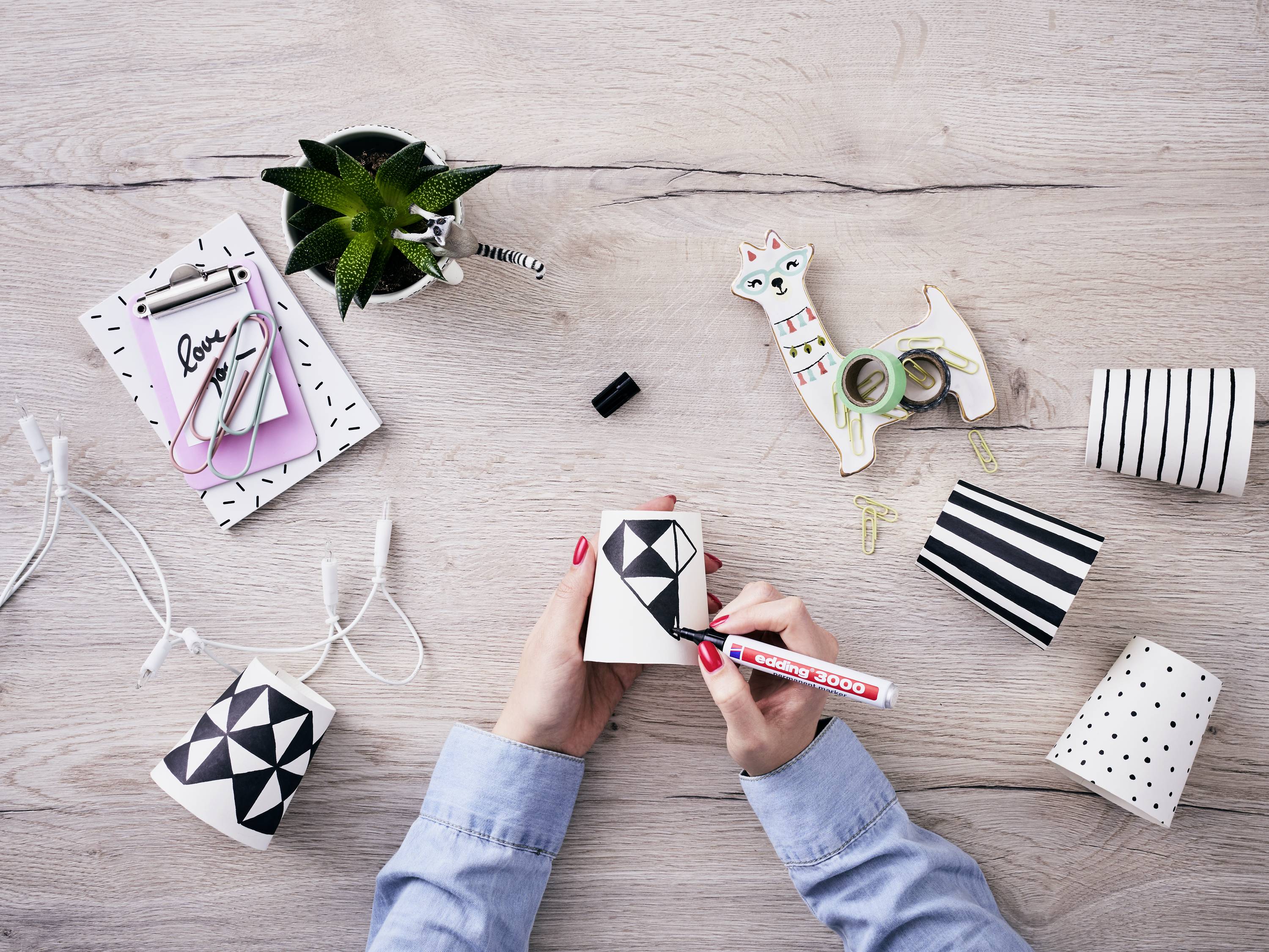 A person is decorating paper cups with geometric patterns on a wooden table, surrounded by paperclips, a notebook, and a plant.