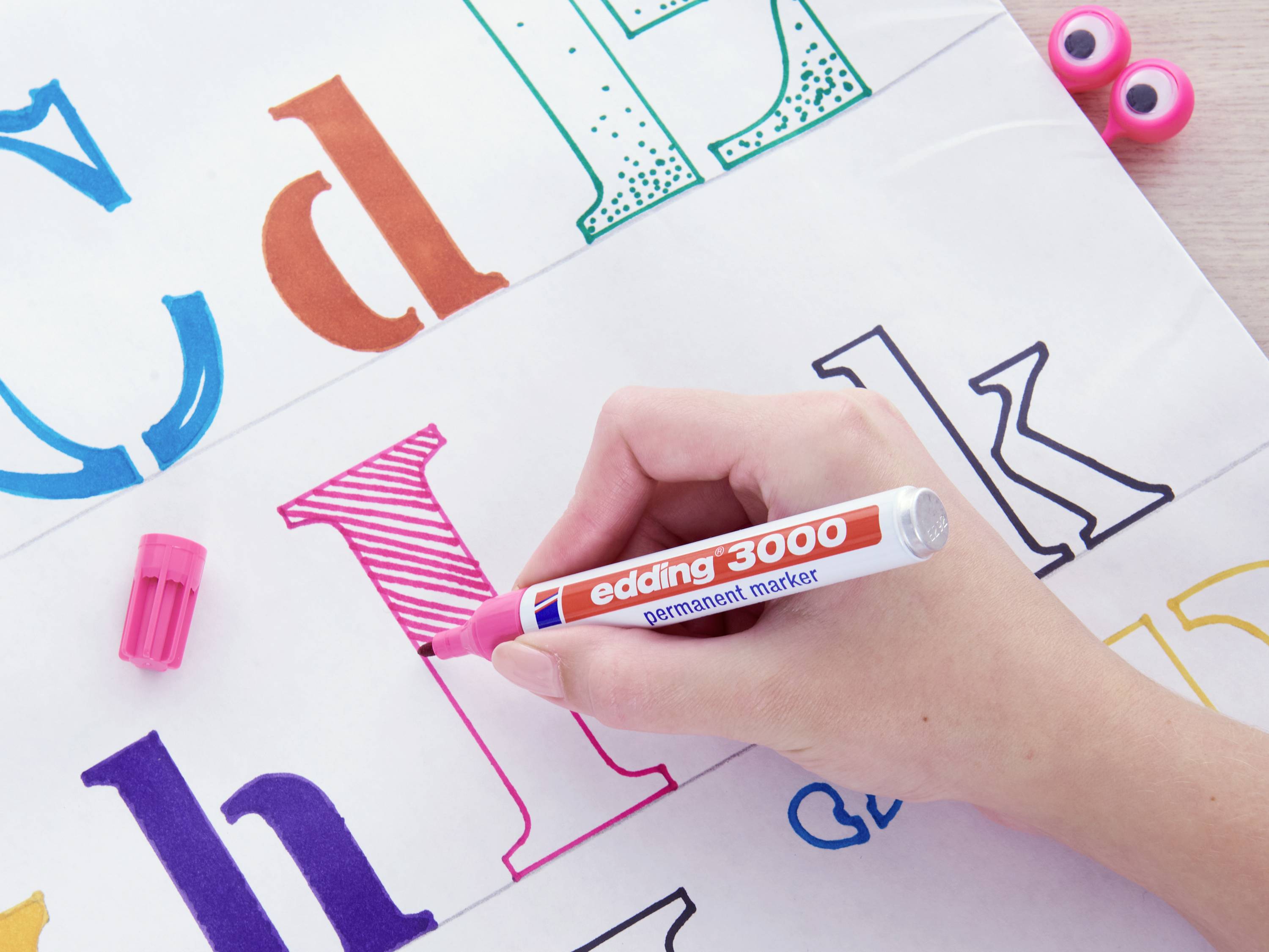 A hand is drawing the letter 'H' with a pink Edding 3000 marker on white paper, surrounded by coloured letters.