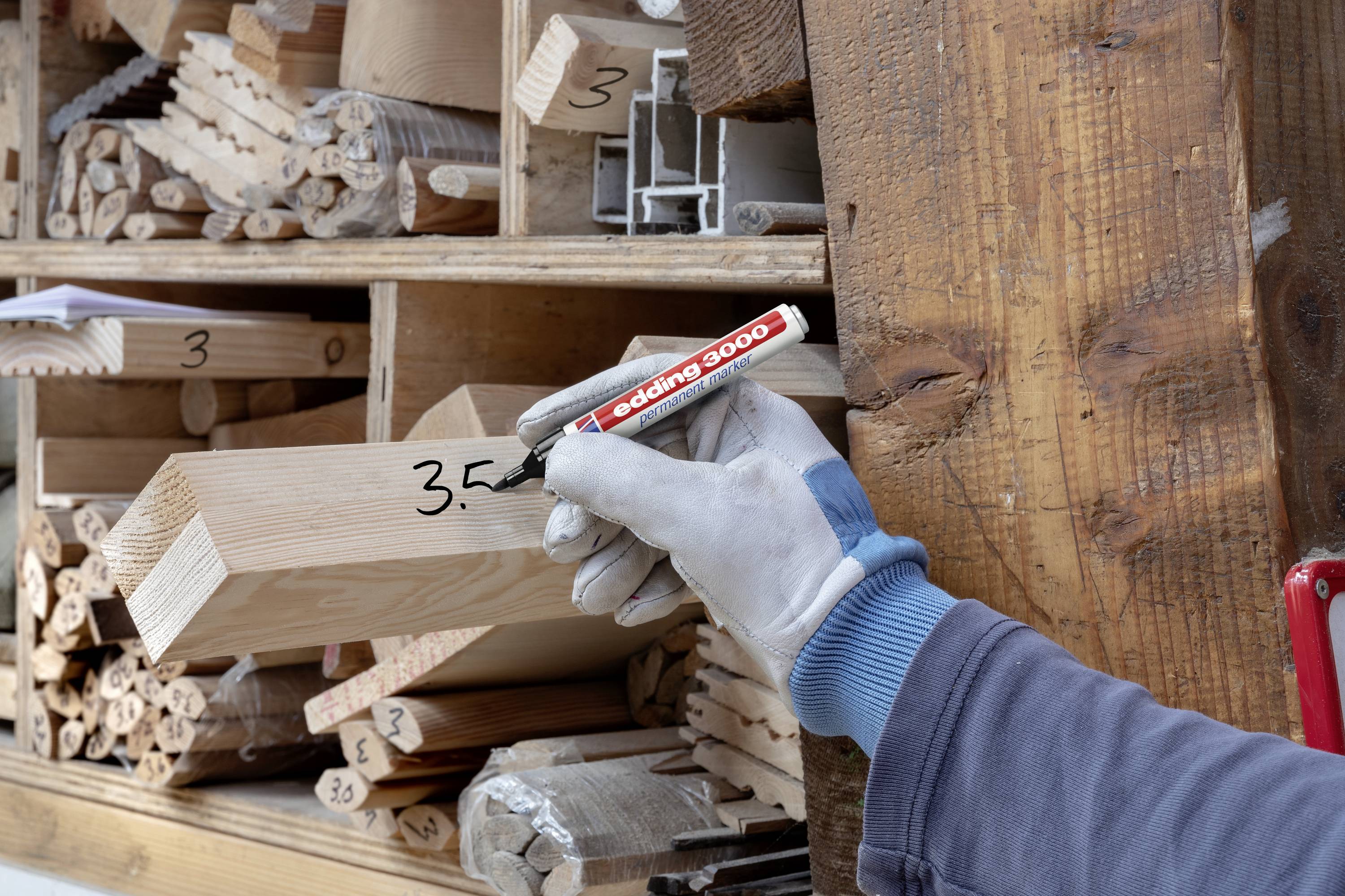 A gloved person is labelling a piece of wood with '3.5' on a shelf full of wooden materials and profiles.