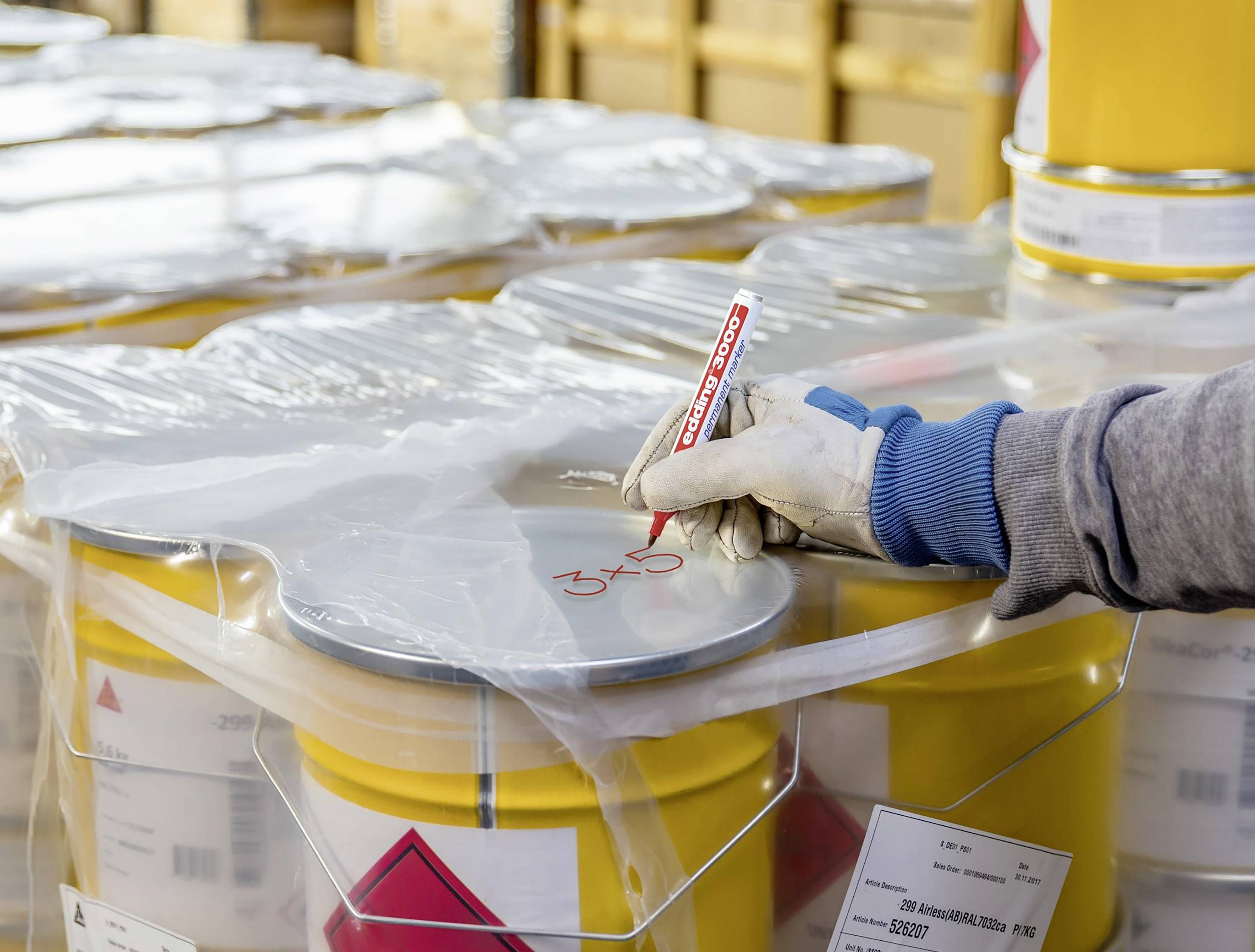 A person is labelling a metal container with a red pen. The container is wrapped in film. Other containers are visible.