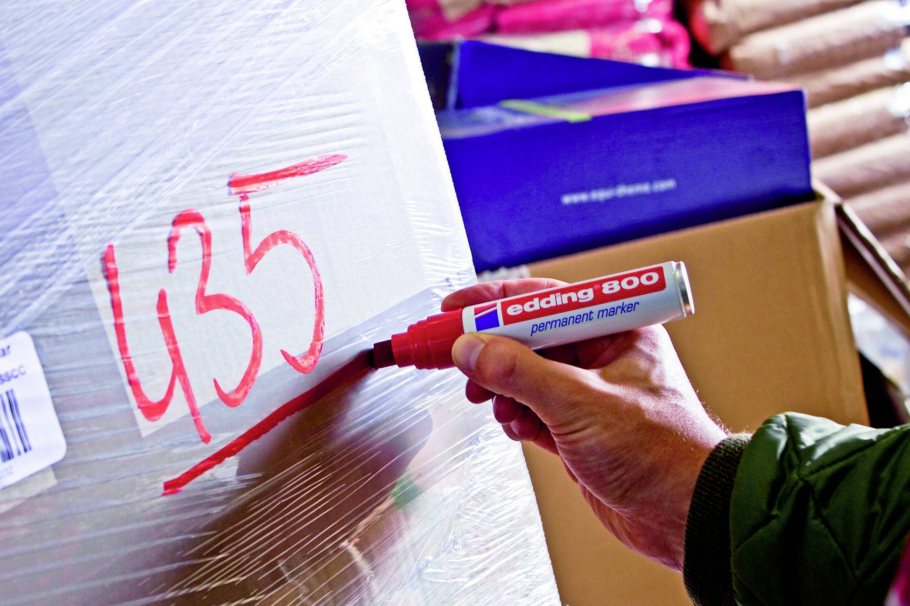 A person is writing the number '435' with a red permanent marker on a wrapped pallet in a warehouse.