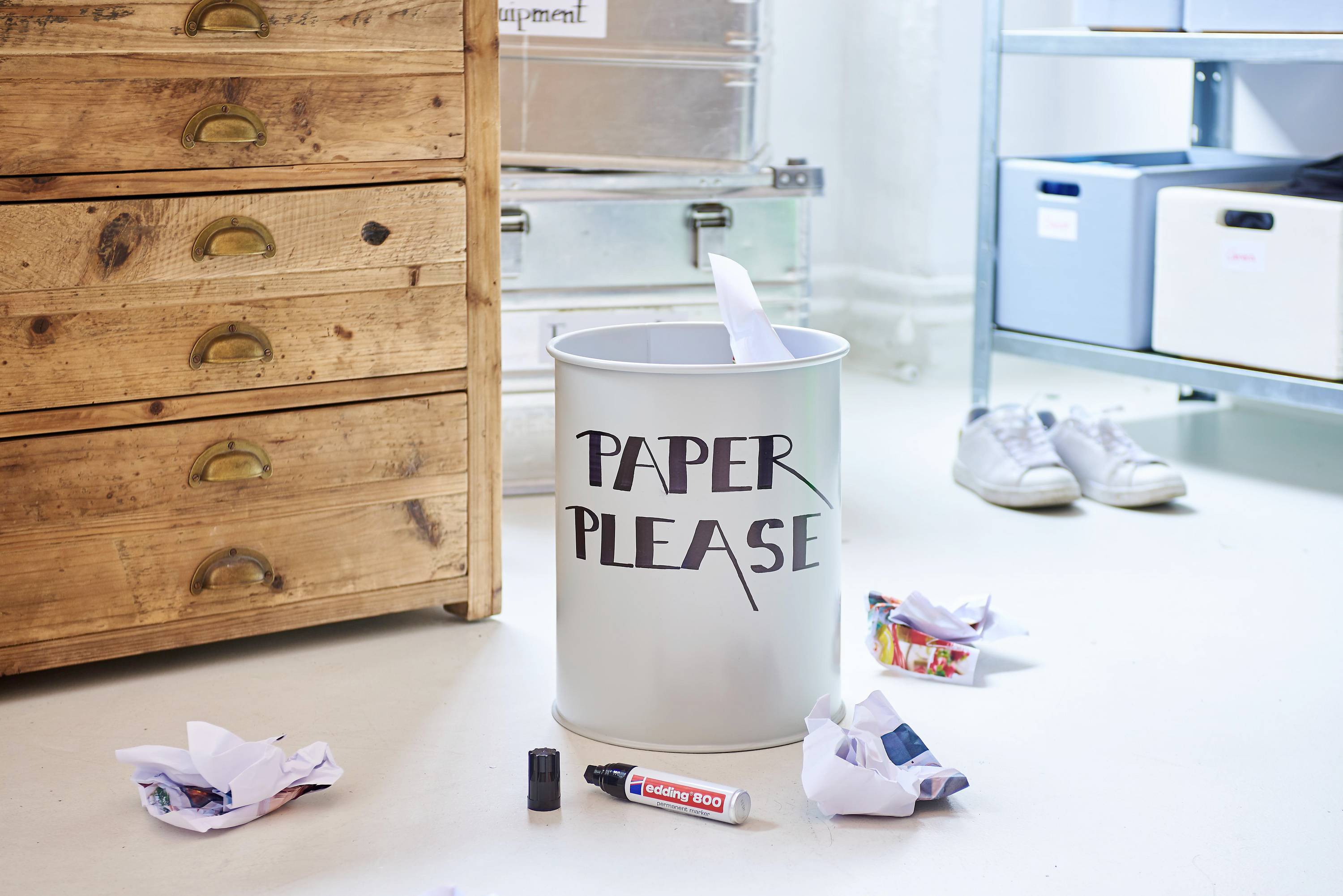 Wastepaper bin labelled 'Paper Please', surrounded by crumpled papers and office supplies scattered on the floor.