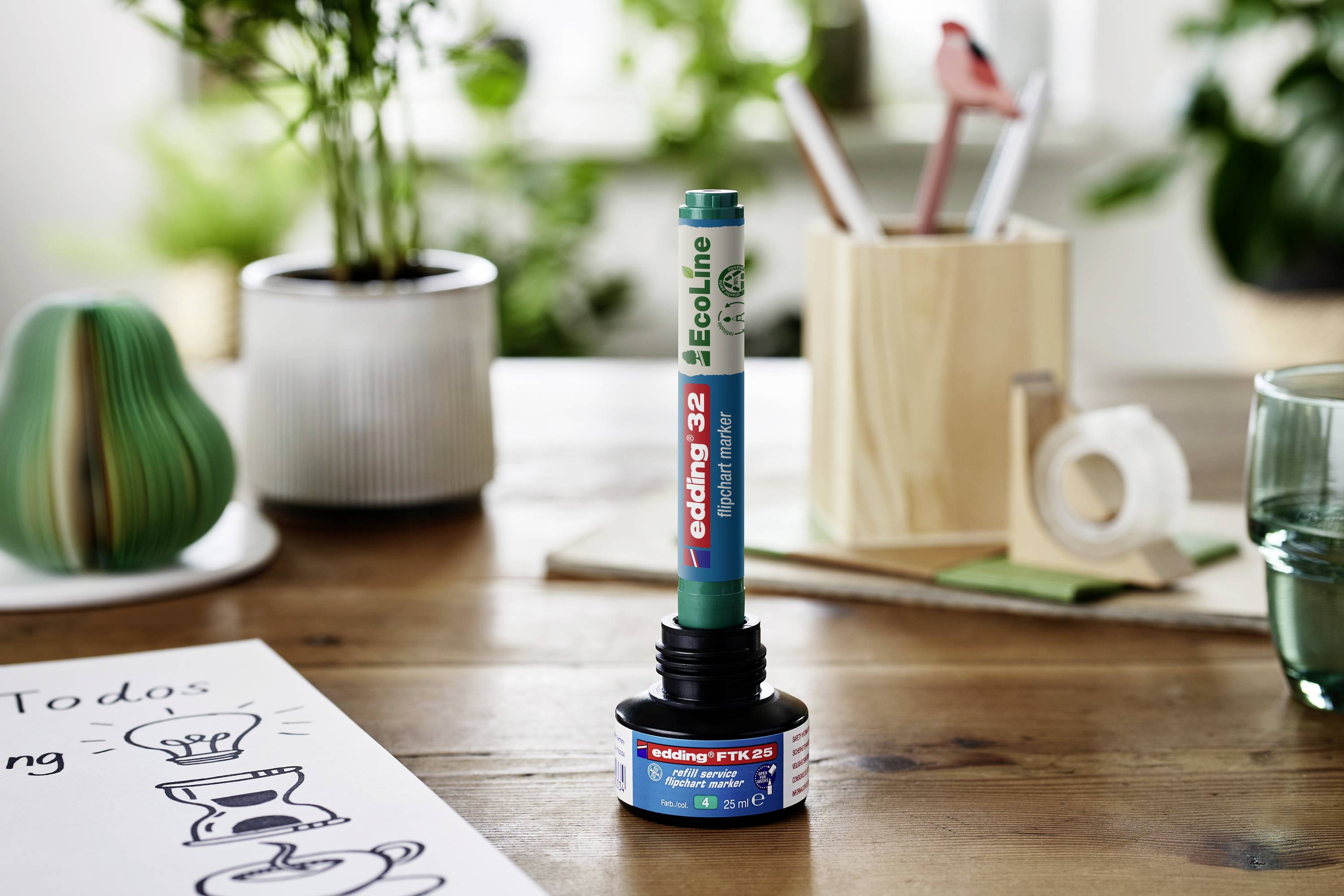 A refillable ink bottle with a green pen, surrounded by plants, a wooden pencil holder, and notepaper on a table.