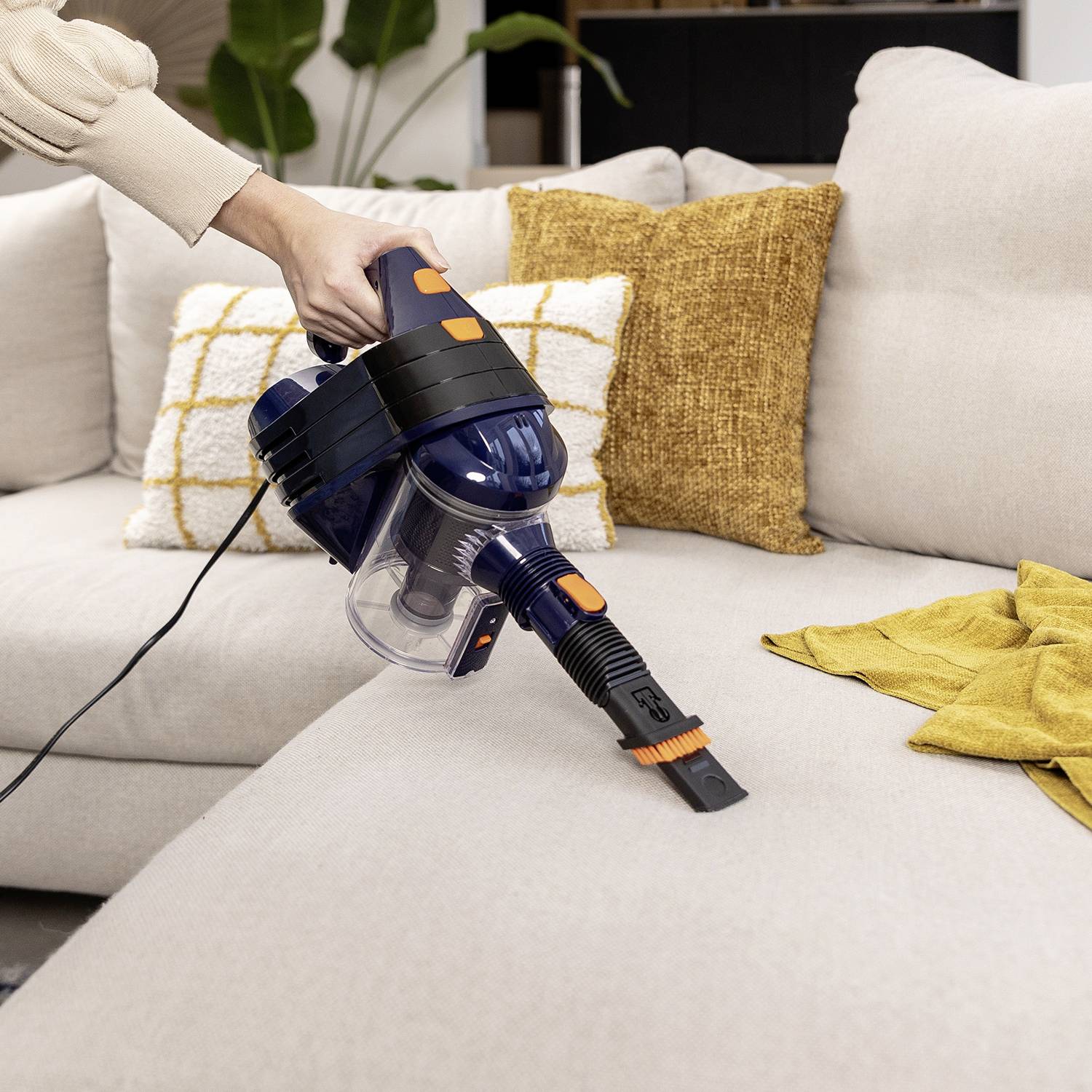 A person is vacuuming a beige sofa with a cordless handheld hoover; cushions can be seen in the background.