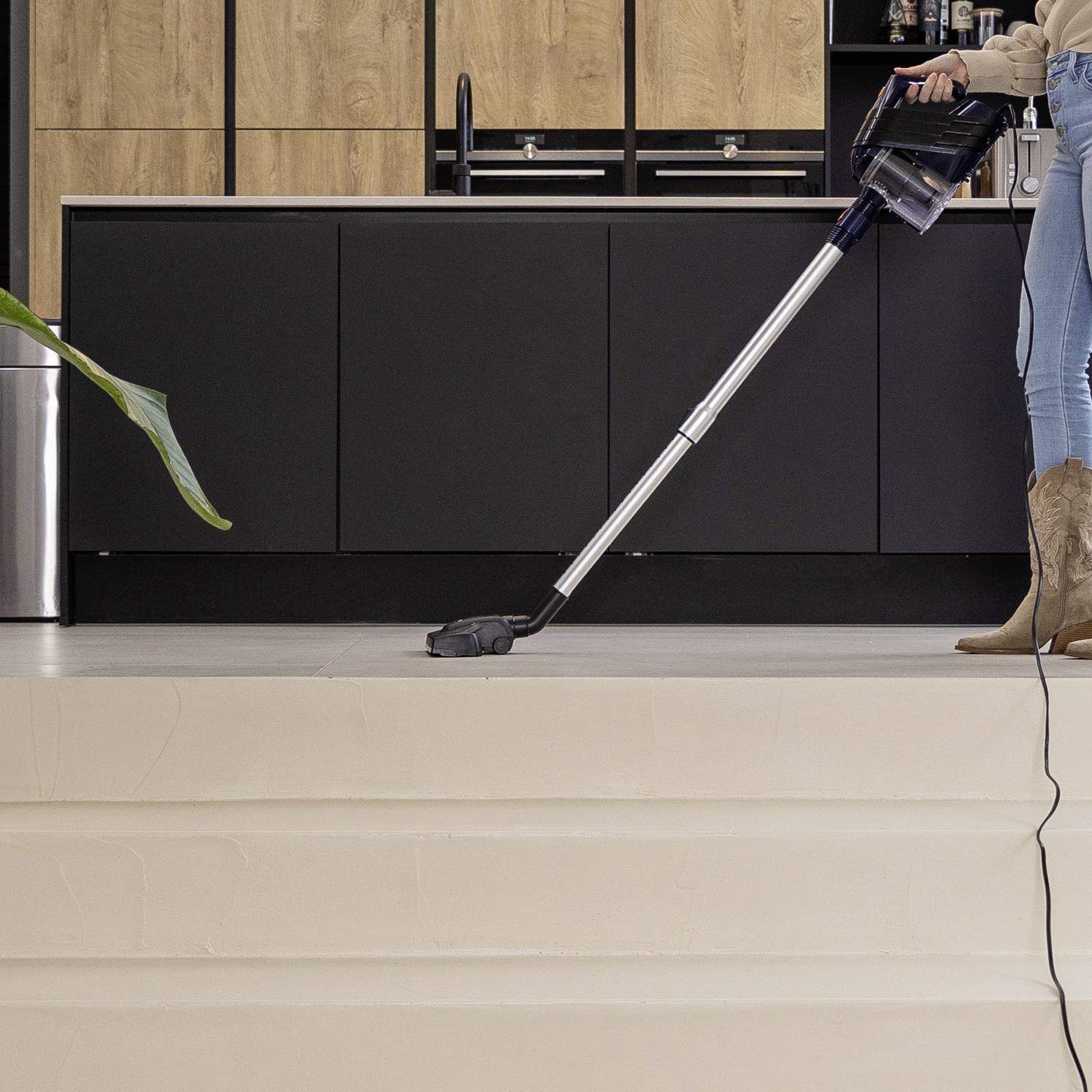 A person is hoovering the floor in a modern kitchen with wooden elements. Kitchen cabinets and a worktop are visible in the background.