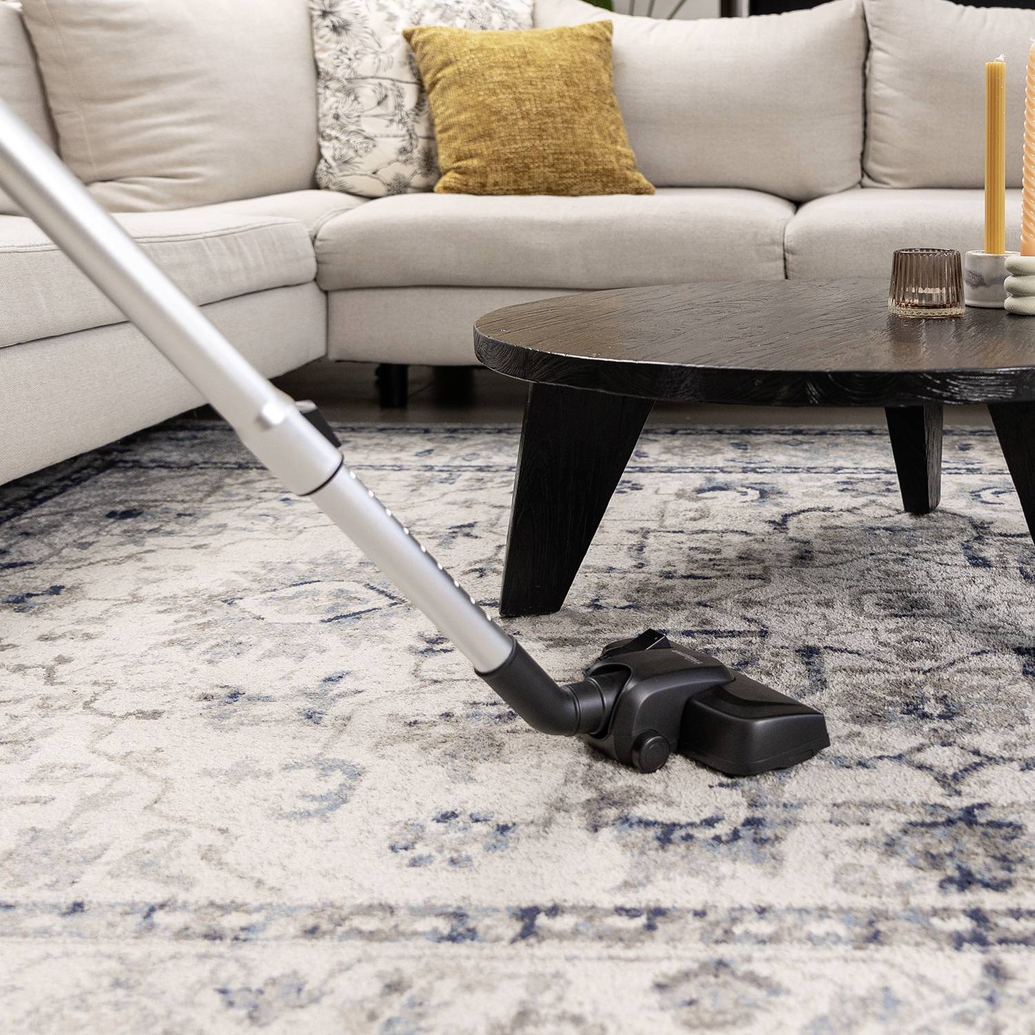A hoover is cleaning a light-coloured carpet in a living room with a beige sofa and a round, dark coffee table.