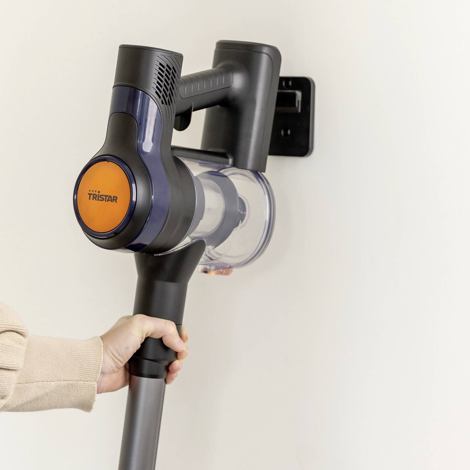 A hand is holding a cordless vacuum cleaner mounted on a wall bracket. The vacuum cleaner is designed with a modern aesthetic.