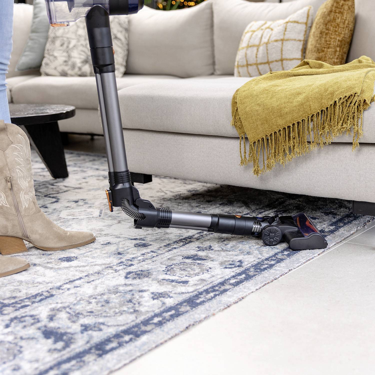 A person is using a cordless vacuum cleaner on a carpet next to a sofa. A yellow cushion and a throw are visible.