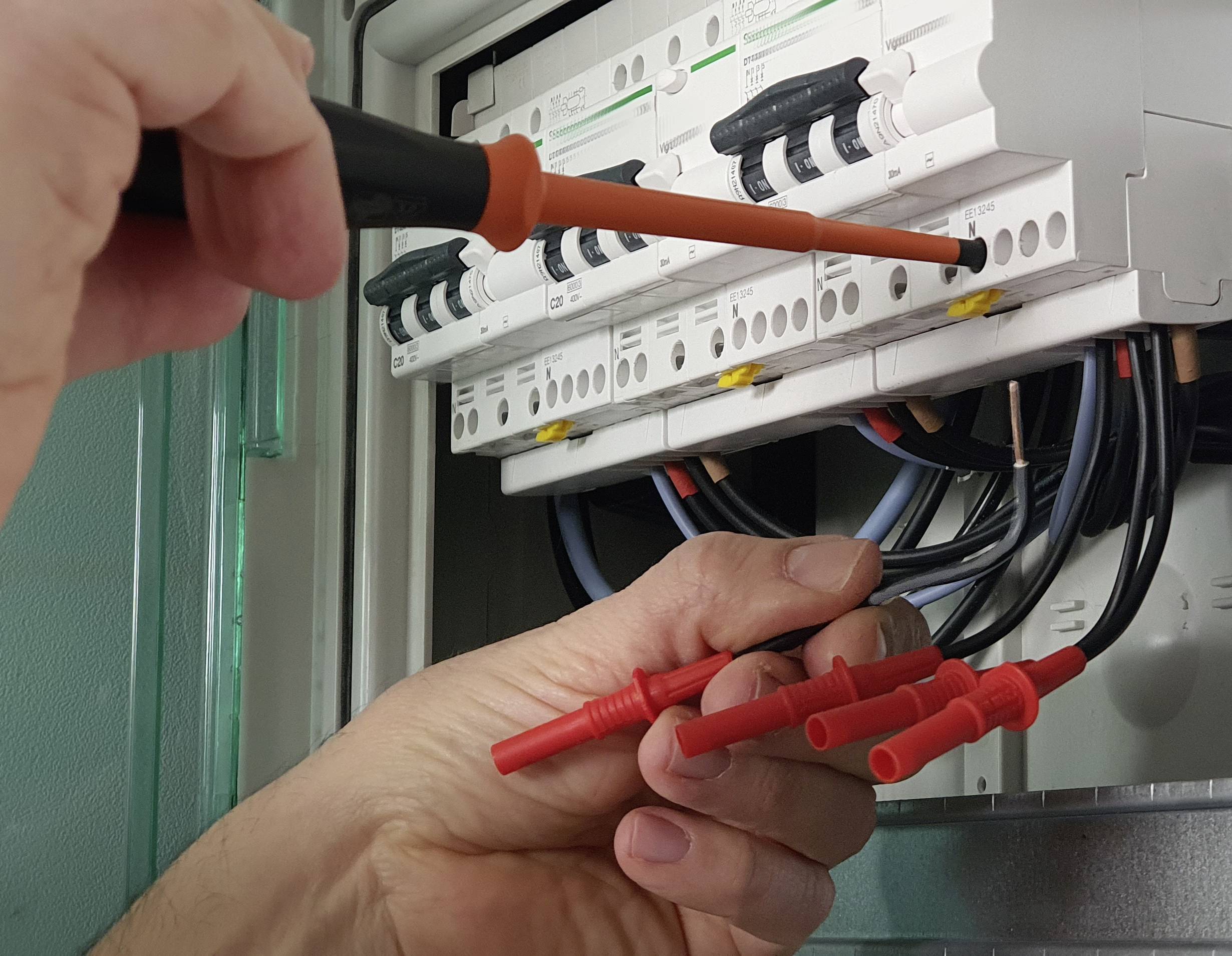 A person is working on a fuse box, checking cables with a screwdriver and test leads.
